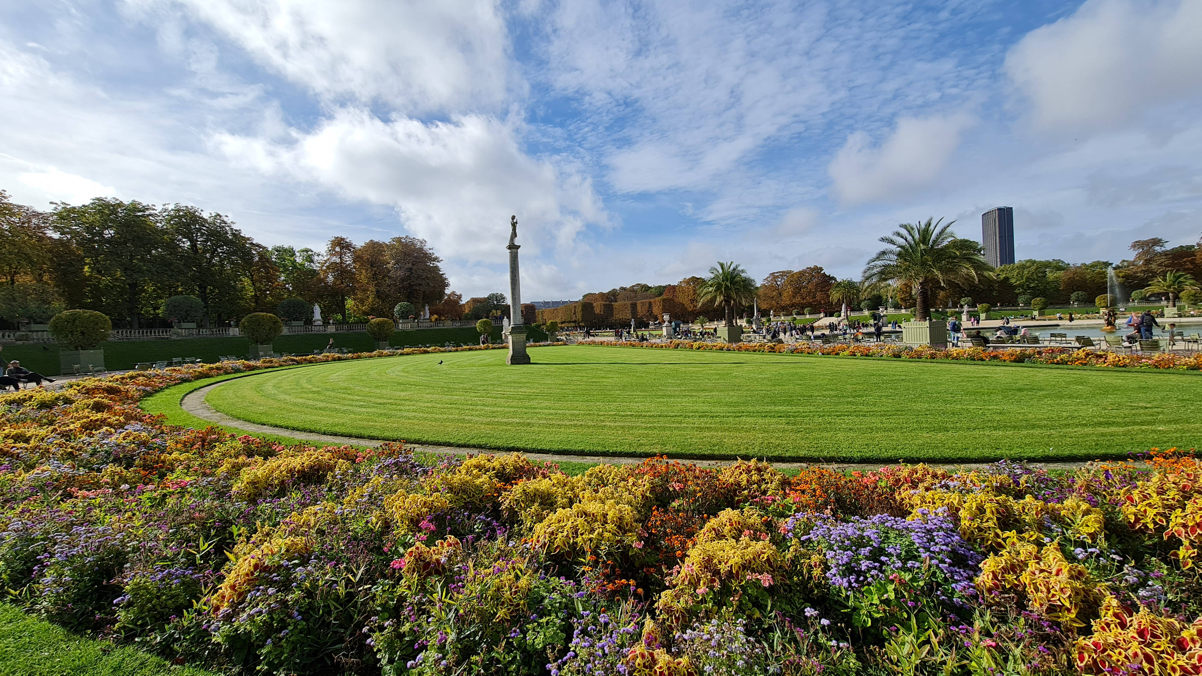 Luxembourg 桌布 壁紙 — 一個充滿活力的花園，中央有圓形草坪，周圍環繞著色彩繽紛的花朵，天空部分多雲。