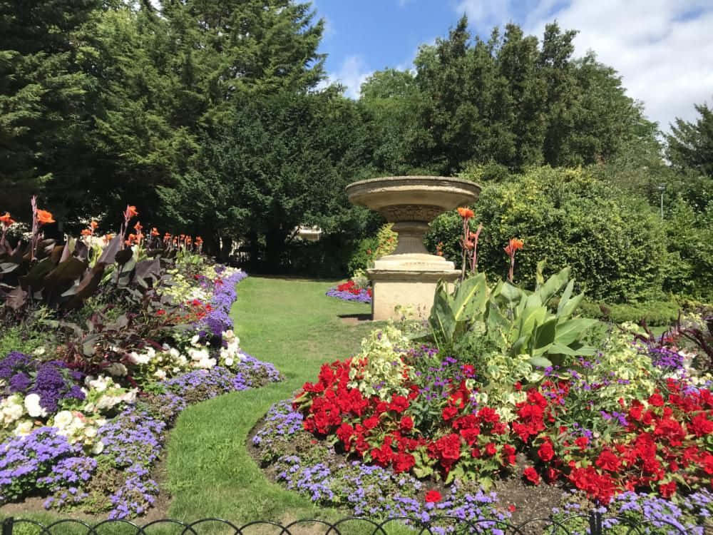 公園 桌布 壁紙 — 一個充滿活力的花園，在明亮的藍天下，石噴泉周圍盛開著紅色、紫色和白色的花朵。