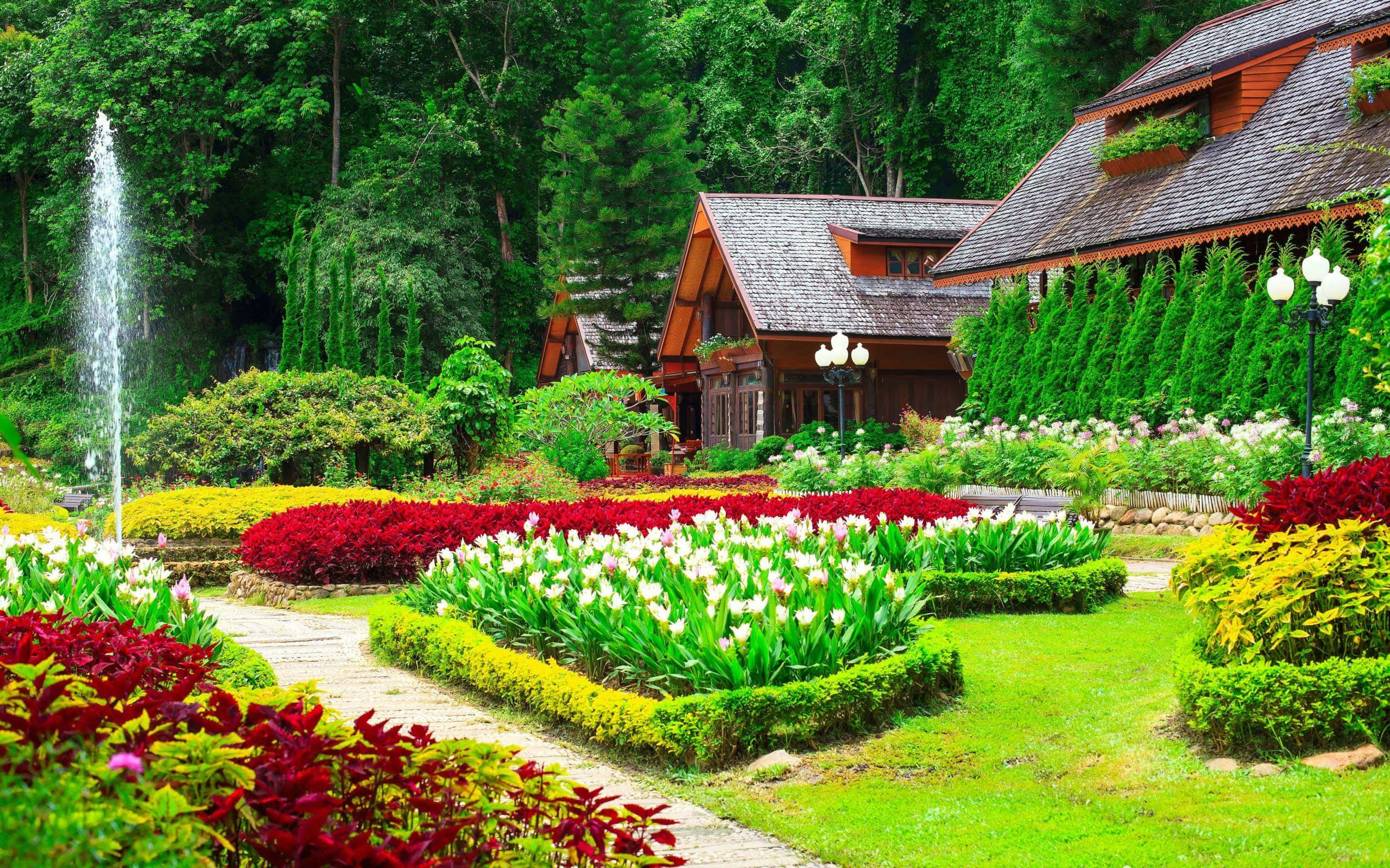 風景 桌布 壁紙 — 一個充滿活力的花園，開滿了紅色、白色和黃色的花朵，環繞著一座木屋，木屋有一個噴泉在茂密的綠色森林中噴水。