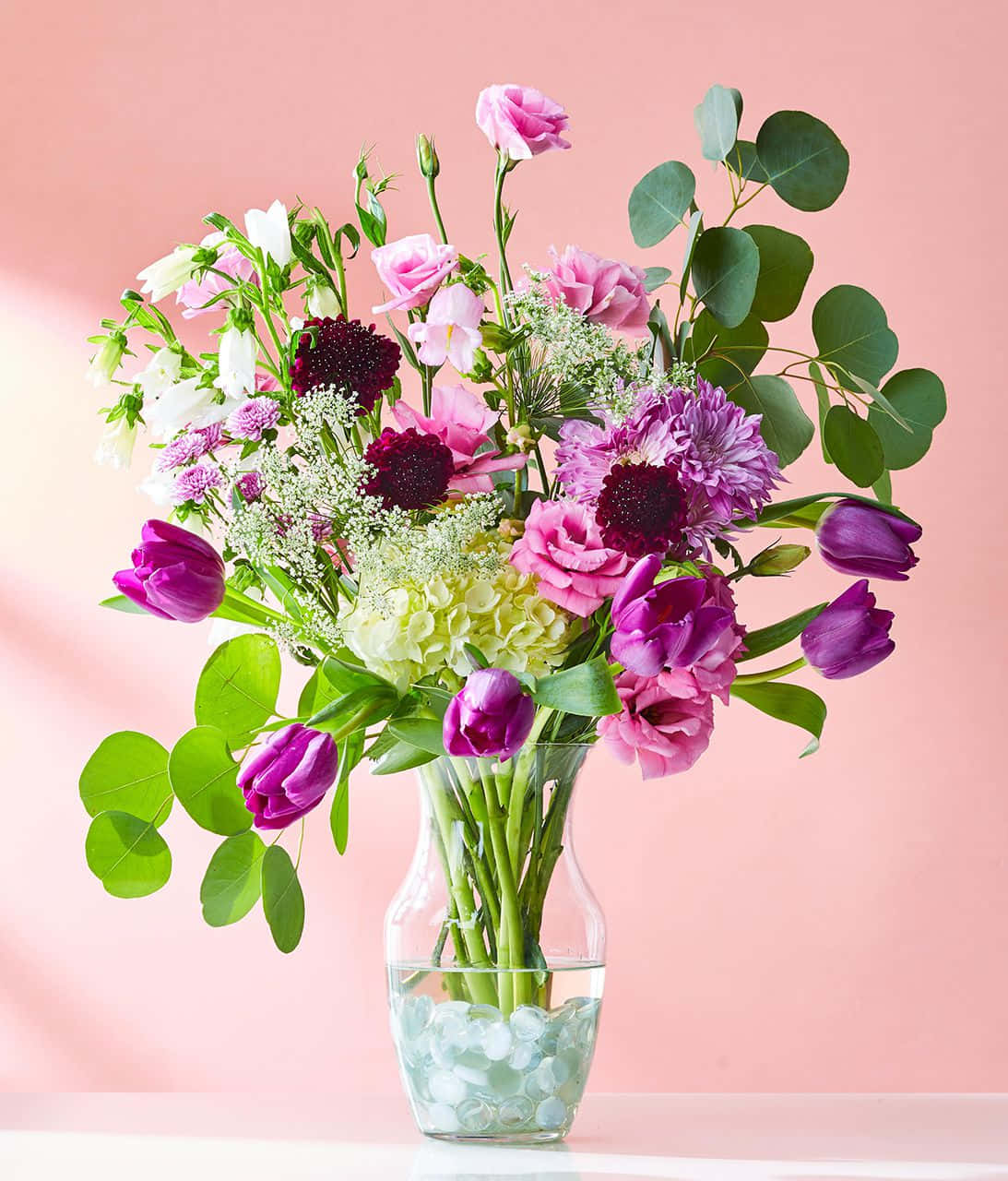 Flowers 桌布 壁紙 — 一個充滿活力的花束，包含粉紅色、紫色和白色的花朵，搭配綠色植物，放在一個裝滿水和鵝卵石的玻璃花瓶中，背景是柔和的粉紅色。
