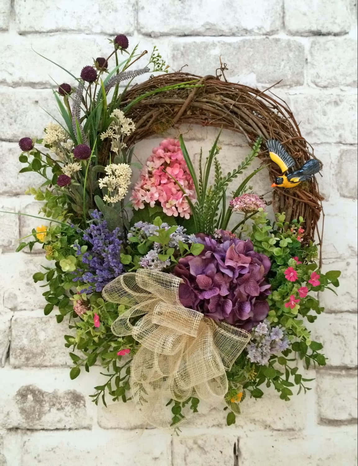 Spring Wreaths 桌布 壁紙 — 一個充滿活力的花環，帶有粉紅色、紫色和白色的花朵、綠色植物和一個黃色小鳥裝飾，掛在白色磚牆上。