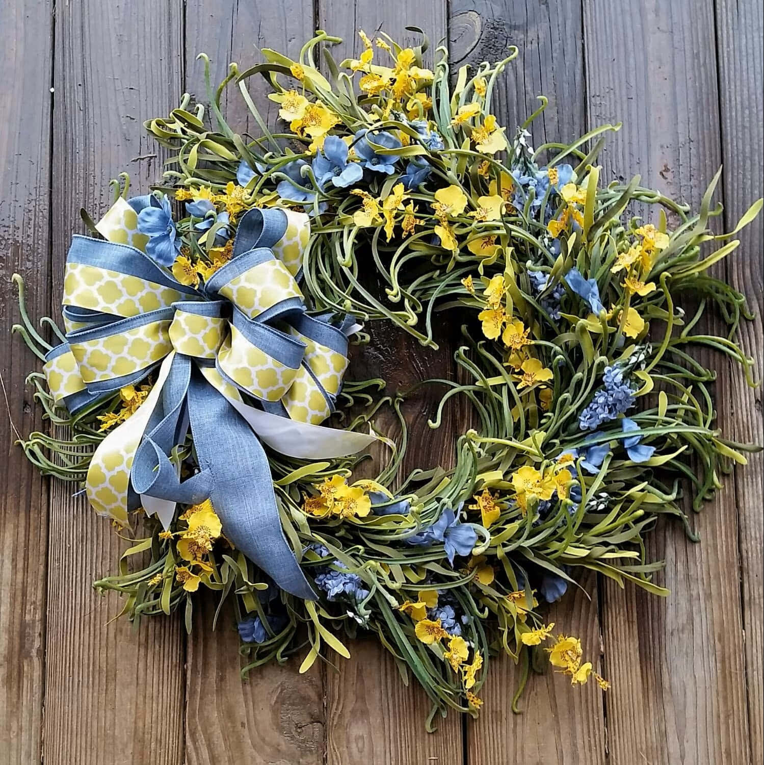 Spring Wreaths 桌布 壁紙 — 一個充滿活力的花環，飾有黃色和藍色的花朵以及圖案蝴蝶結，放在風化的木板上。