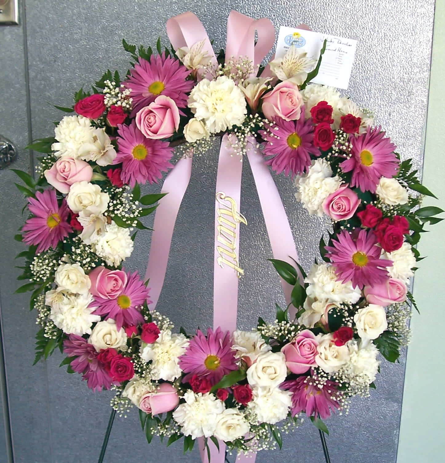 Funeral Flower Arrangements 桌布 壁紙 — 一個充滿活力的花環，點綴著粉紅玫瑰、雛菊和白色康乃馨，掛在灰色的門上，帶有粉紅絲帶，上面寫著'Faith'。