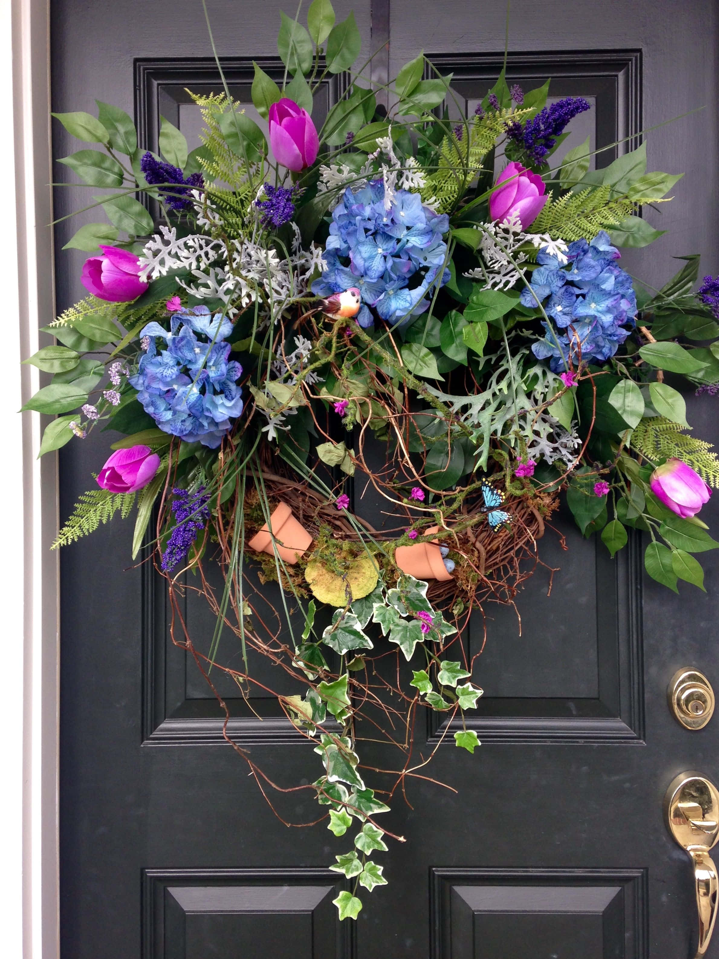 Spring Wreaths 桌布 壁紙 — 一個充滿活力的花環，點綴著藍色繡球花、粉紅色鬱金香和紫色花朵，裝飾在深色的大門上。