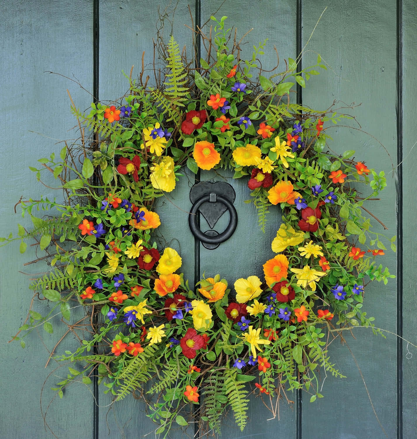 Spring Wreaths 桌布 壁紙 — 一個充滿活力的花環，點綴著黃色、橙色、紅色和紫色的花朵以及綠色蕨類植物，裝飾在一扇帶有黑色金屬門環的風化青綠色木門上。