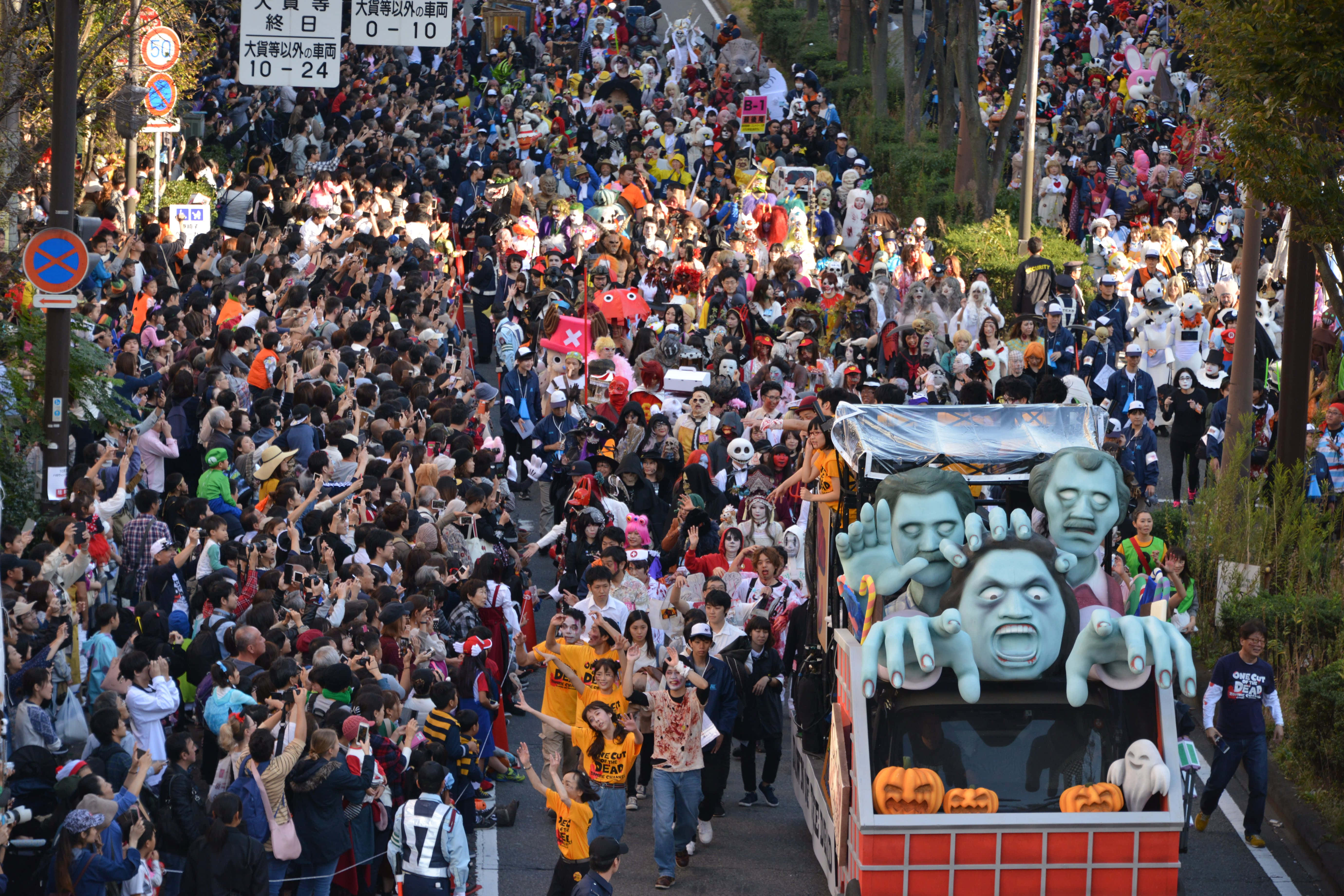 Halloween Parades 桌布 壁紙 — 一個充滿活力的萬聖節遊行，擠滿了樹木成蔭的街道，盛裝打扮的人群歡呼著，一輛以殭屍為主題、裝飾著發光南瓜的花車緩緩駛過。