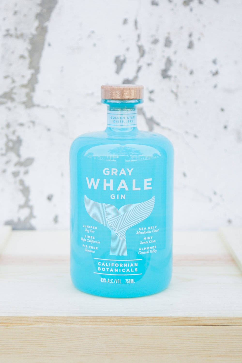 alcohol 桌布 壁紙 — 一個充滿活力的藍綠色Gray Whale Gin酒瓶，帶有木質瓶蓋，置於淺色木質表面中央，背景為有紋理的白色牆壁。