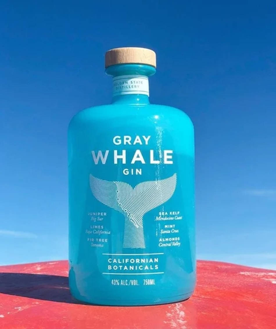 bottle 桌布 壁紙 — 一個充滿活力的藍綠色Gray Whale Gin酒瓶，帶有木蓋，放在紅色表面上，背景是清澈的藍天。