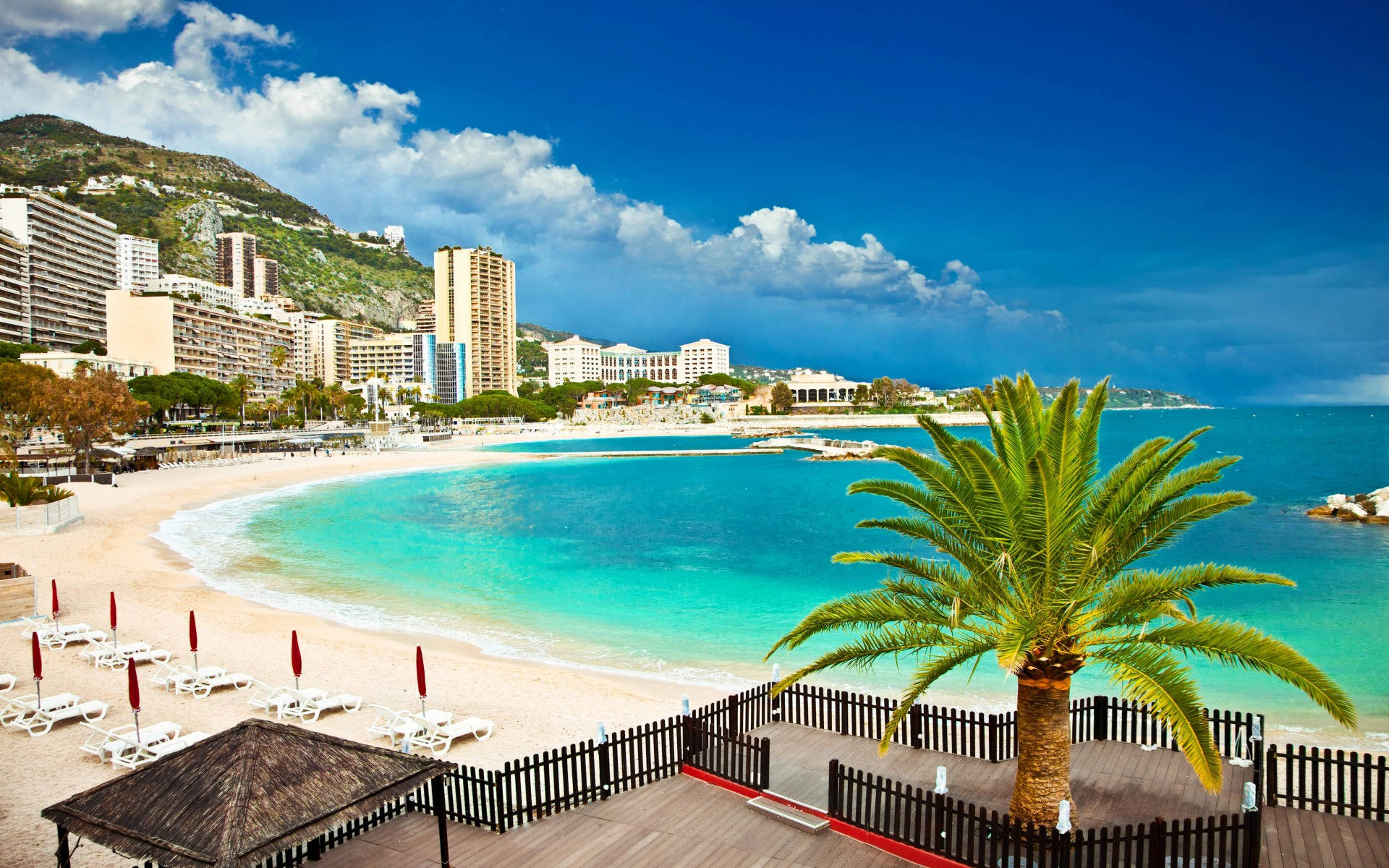 Monaco 桌布 壁紙 — 一個充滿活力的藍綠色海灣蜿蜒環繞著一個沙灘，沙灘上排列著白色躺椅和紅色遮陽傘，上方是晴朗的藍天和蓬鬆的白雲。