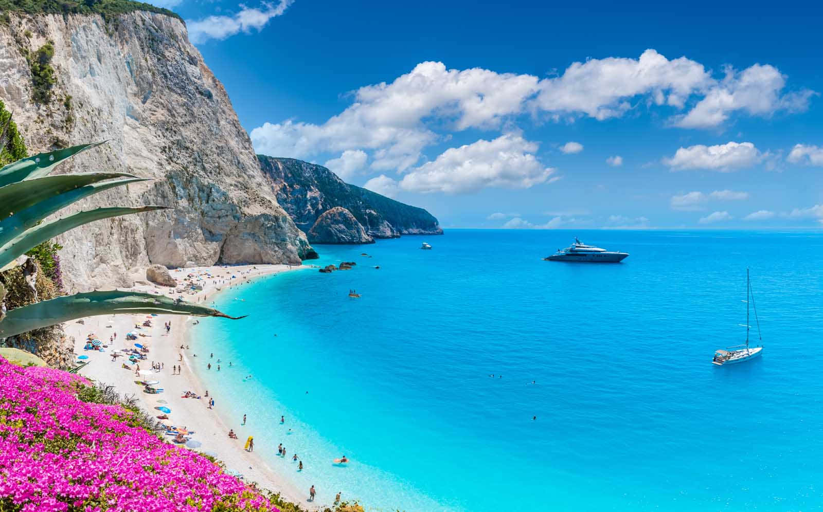 Greek Beach 桌布 壁紙 — 一個充滿活力的藍綠色海灣蜿蜒環繞著一個白色沙灘，沙灘上擠滿了享受日光浴的人們，上方是點綴著蓬鬆雲朵的明亮藍天。