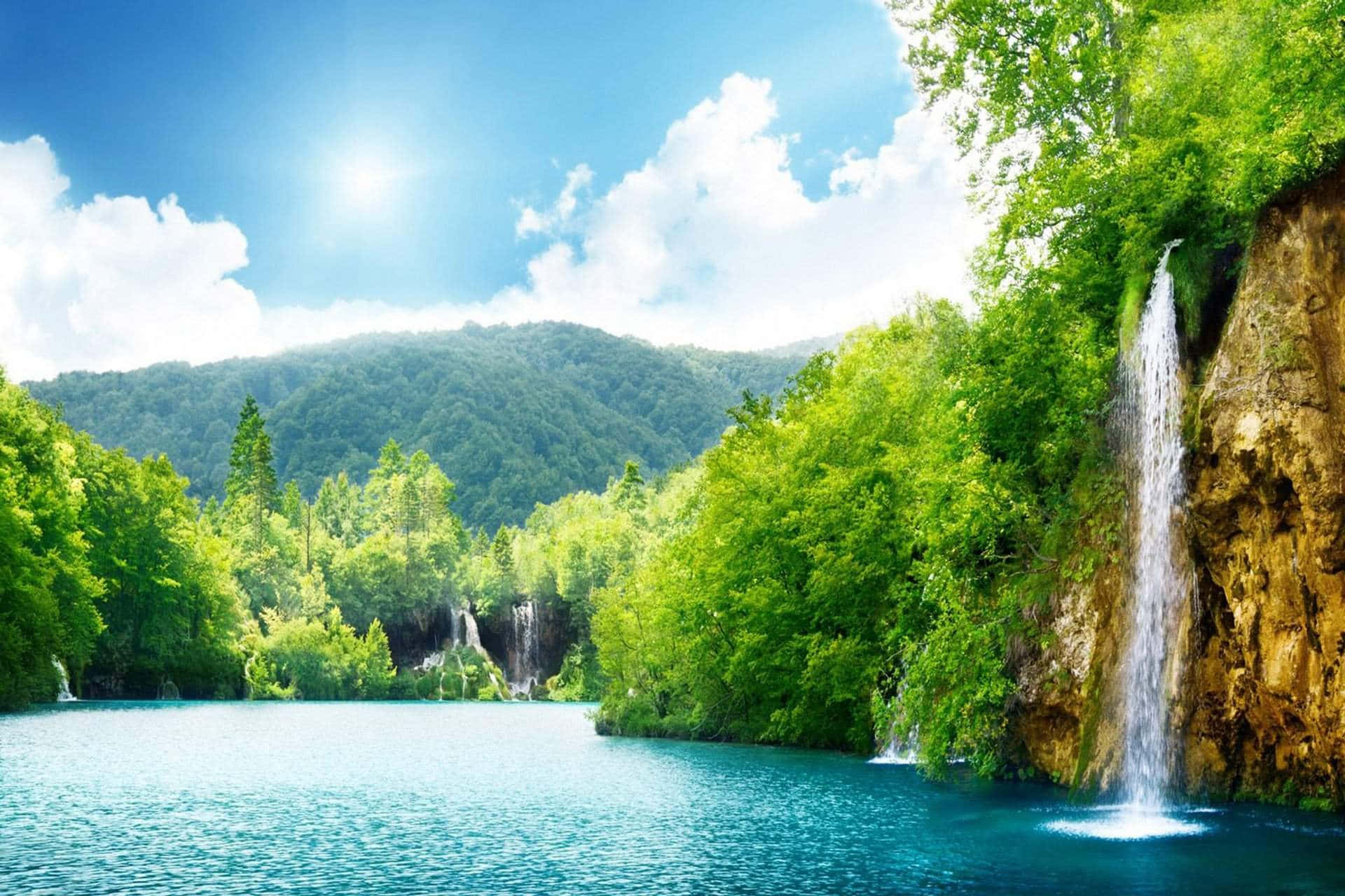 Beautiful View 桌布 壁紙 — 一個充滿活力的藍綠色湖泊，周圍環繞著茂密的綠色森林和層疊瀑布，在明亮的藍天下有蓬鬆的雲朵。
