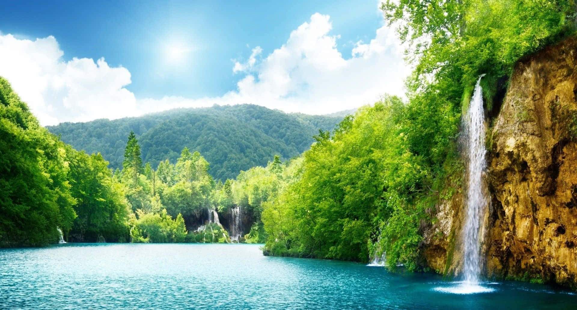 1920X1080 桌布 壁紙 — 一個充滿活力的藍綠色湖泊，周圍環繞著茂密的綠色森林，瀑布在明亮的藍天下從岩石懸崖上傾瀉而下。
