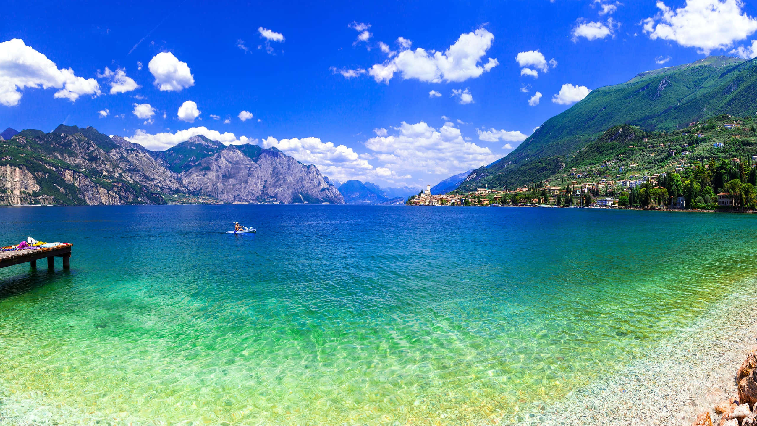 Lake 桌布 壁紙 — 一個充滿活力的藍綠色湖泊，坐落在鬱鬱蔥蔥的綠色山脈之間，上方是明亮的藍天和蓬鬆的白雲。