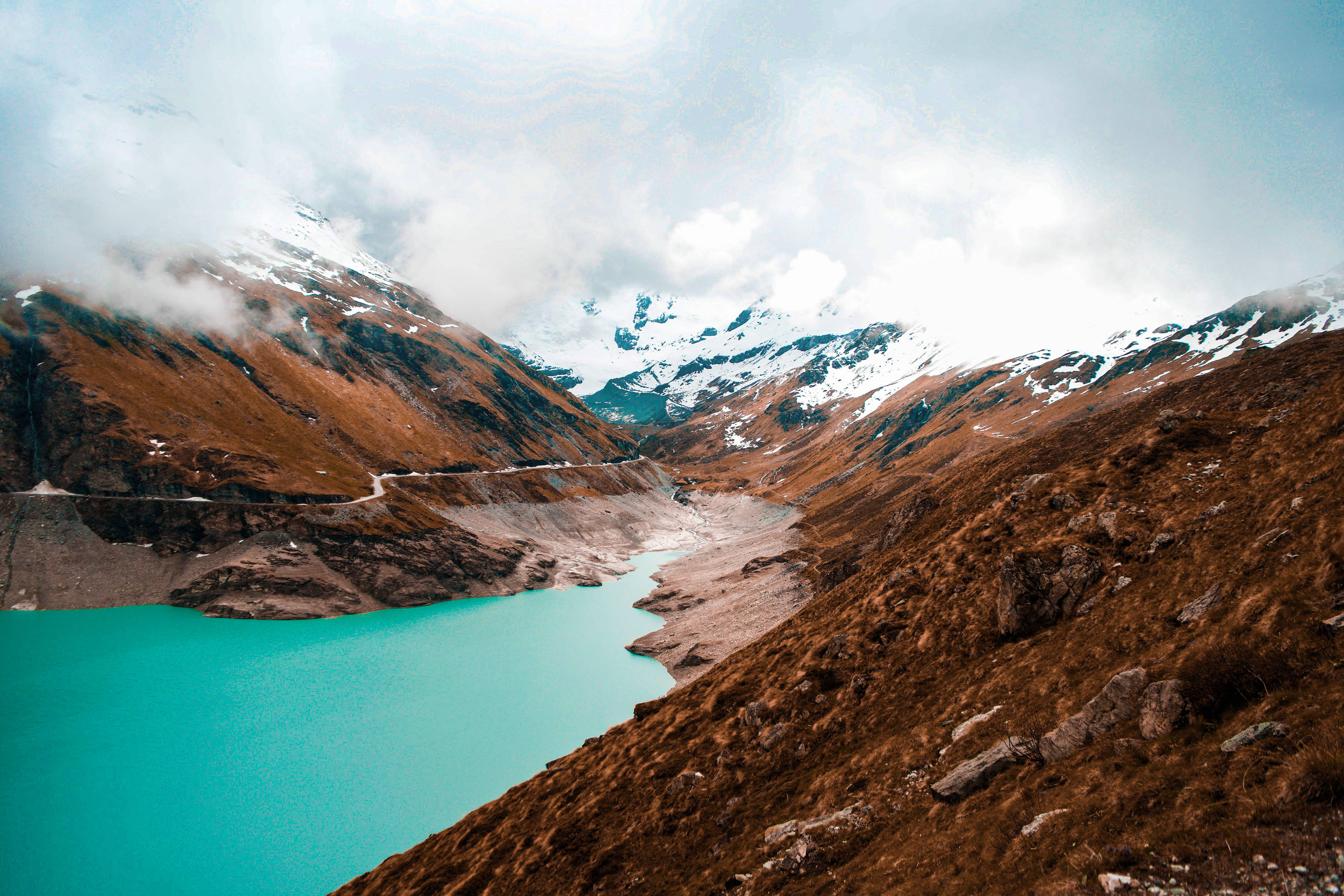 Lake 桌布 壁紙 — 一個充滿活力的藍綠色湖泊，坐落在崎嶇的棕色山脈中，山頂積雪，雲霧繚繞。