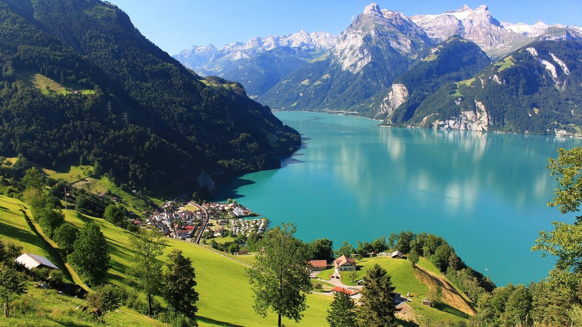 Alps 桌布 壁紙 — 一個充滿活力的藍綠色湖泊，坐落在鬱鬱蔥蔥的綠色山丘和白雪覆蓋的山脈之間，天空清澈湛藍。