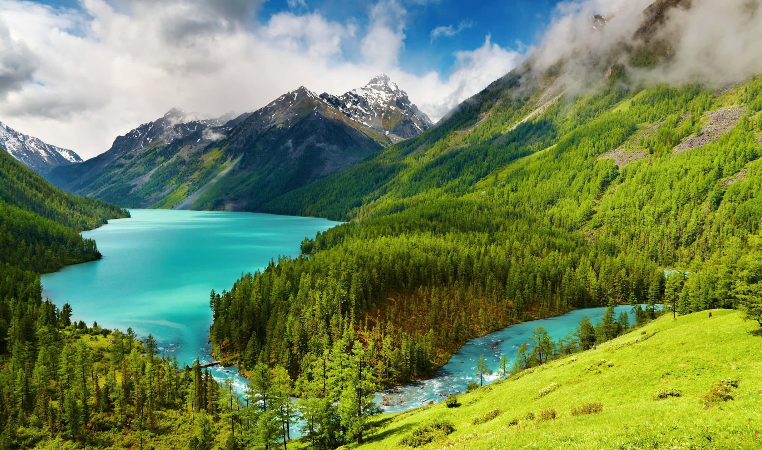 2440x1440 Nature 桌布 壁紙 — 一個充滿活力的藍綠色湖泊，坐落在鬱鬱蔥蔥的綠色山脈中，山頂積雪，天空多雲，藍天白雲。