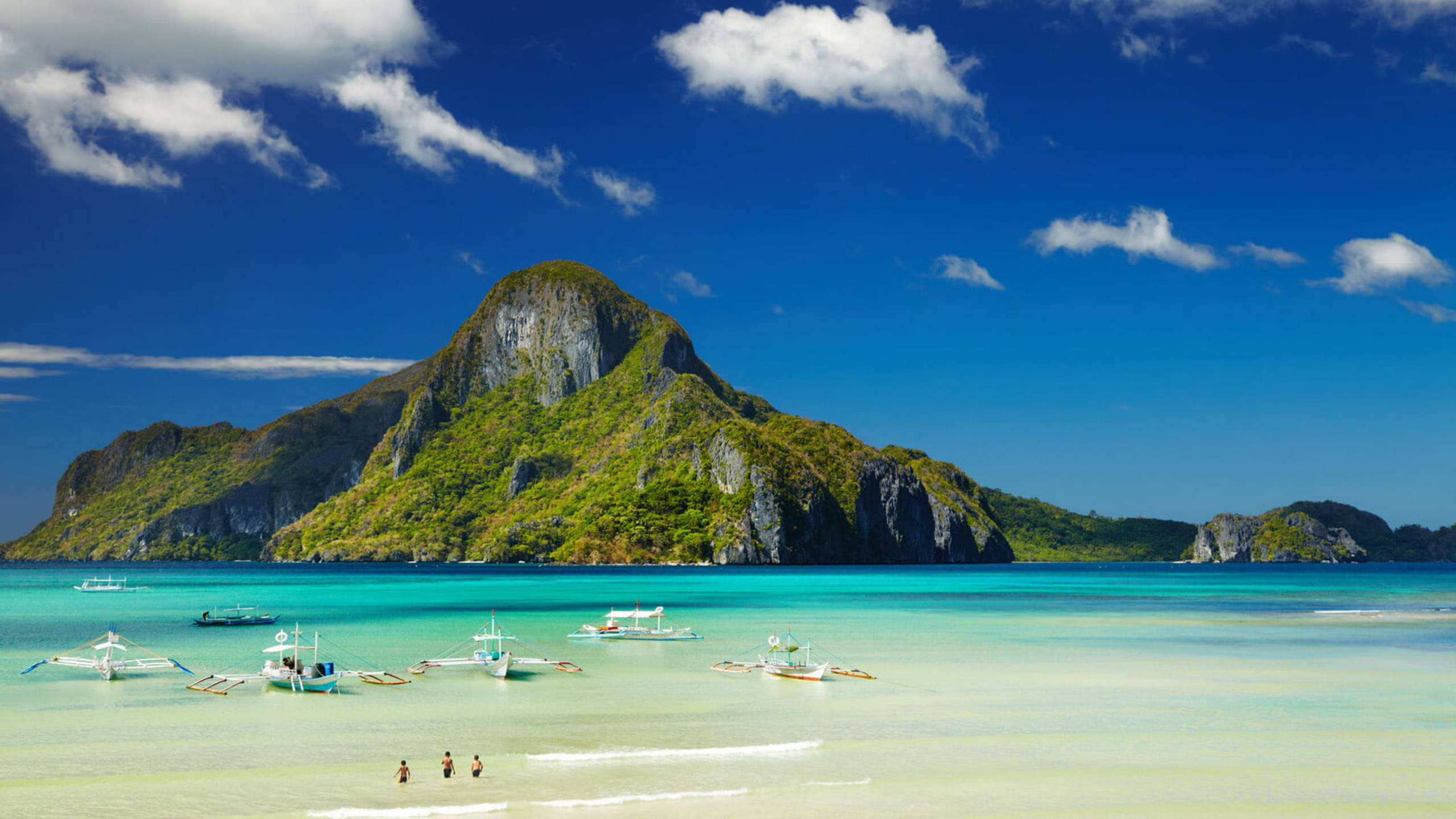 White Beach 桌布 壁紙 — 一個充滿活力的藍綠色潟湖，傳統船隻和小人物靠近岸邊，被茂密的綠色喀斯特島嶼框住，在明亮的藍天下有散落的雲朵。