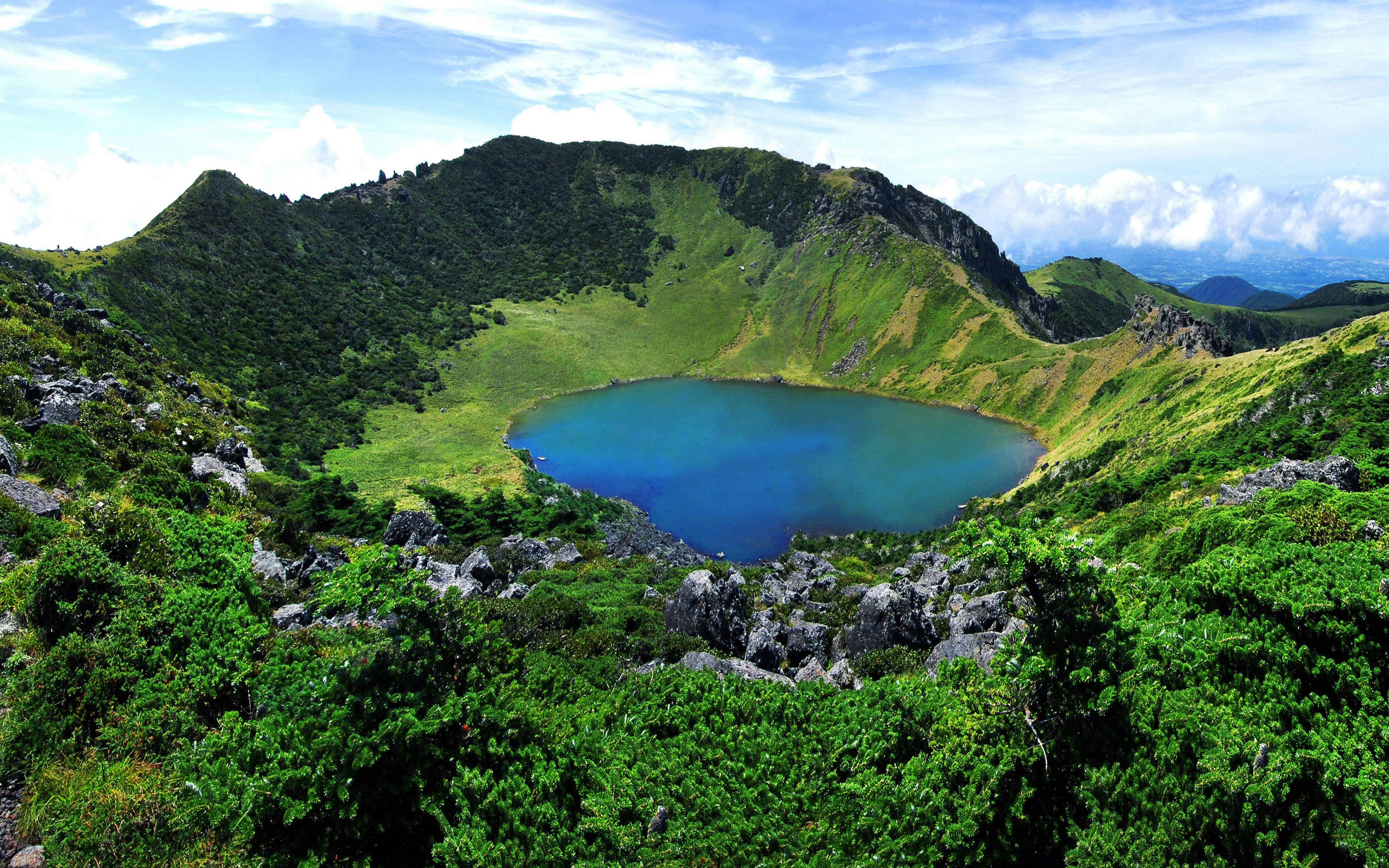 Lake 桌布 壁紙 — 一個充滿活力的藍綠色火山口湖，坐落在鬱鬱蔥蔥的綠色山谷中，上方是明亮的藍天，散佈著幾朵雲。