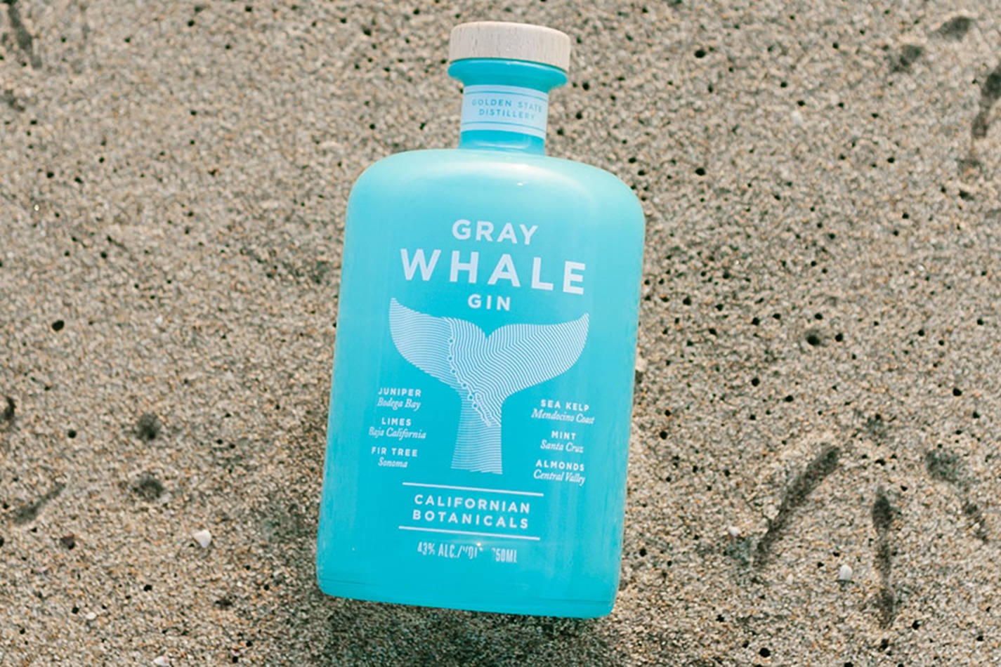 bottle 桌布 壁紙 — 一個充滿活力的藍綠色琴酒瓶，帶有鯨魚尾巴設計，放置在紋理灰色沙子上。