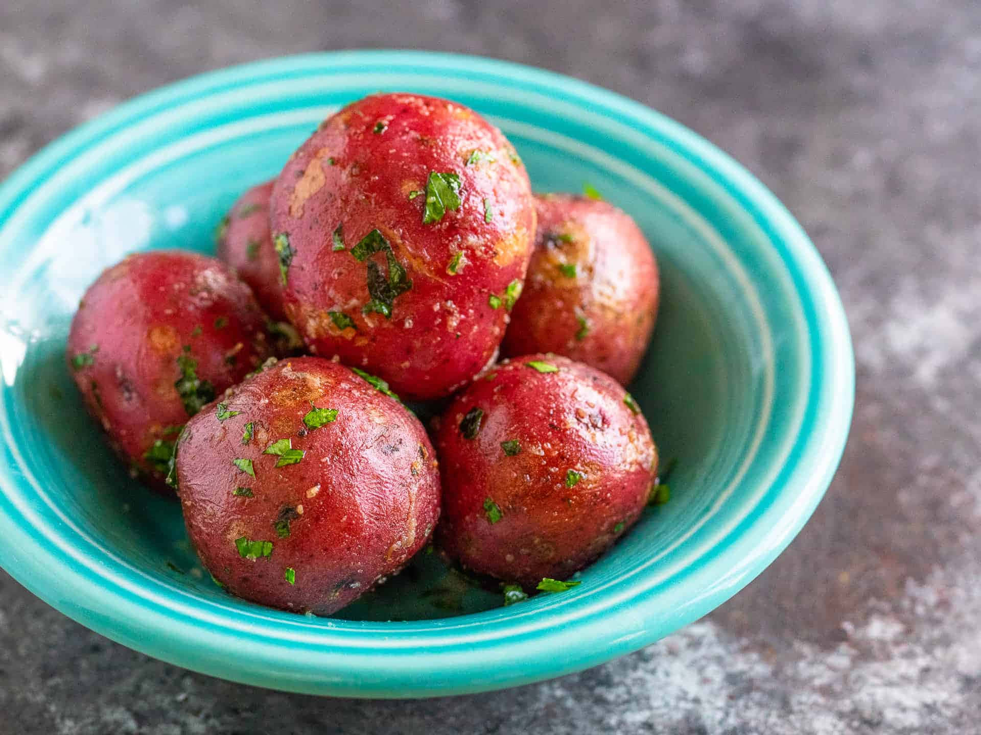 Red Potatoes 桌布 壁紙 — 一個充滿活力的藍綠色碗裡盛著光澤的紅色馬鈴薯，配上新鮮的綠色香草，背景是帶有紋理的灰色。