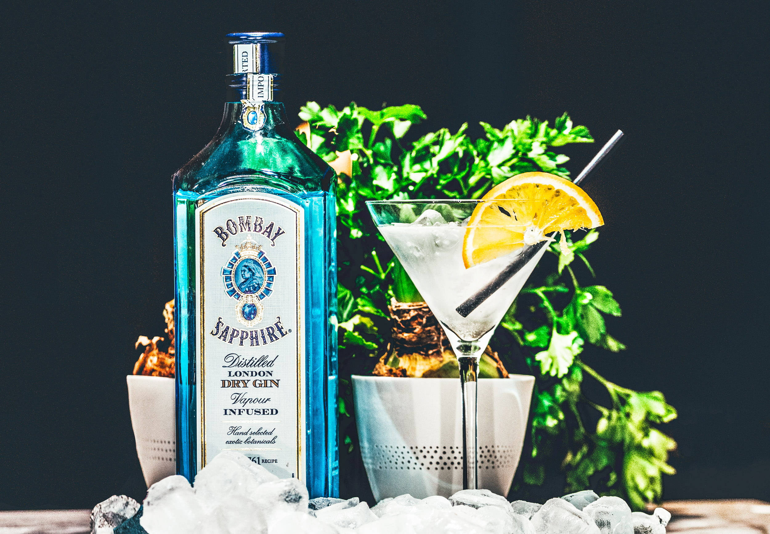 Wine 桌布 壁紙 — 一個充滿活力的藍色 Bombay Sapphire 琴酒瓶旁邊放著一個裝飾有橙片和冰塊的馬丁尼酒杯，背景是深色和綠色植物。