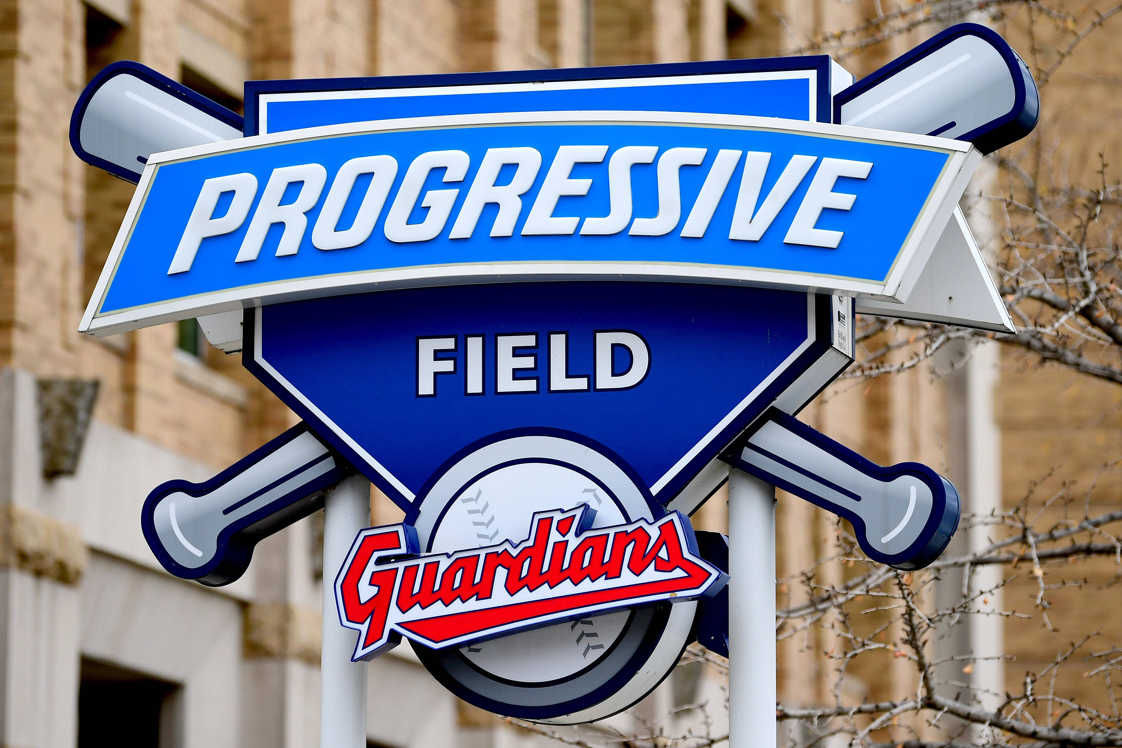 Cleveland Guardians 桌布 壁紙 — 一個充滿活力的藍色 Progressive Field 標誌，帶有交叉的球棒和 Guardians 標誌，以光禿的樹枝和磚砌建築為背景。