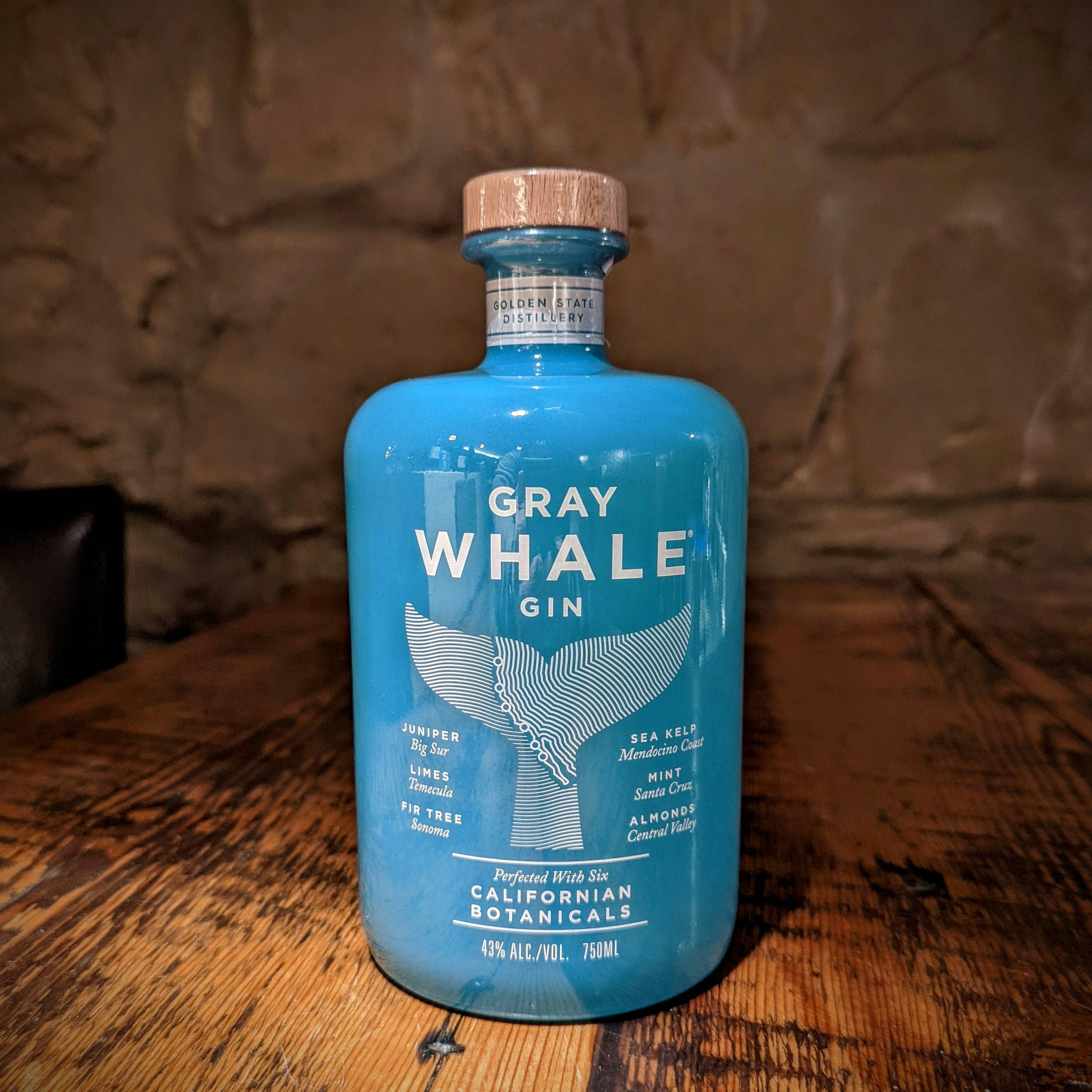 alcohol 桌布 壁紙 — 一個充滿活力的藍色Gray Whale Gin酒瓶，帶有鯨魚尾巴設計，放在一個質樸的木桌上，背景是石牆。
