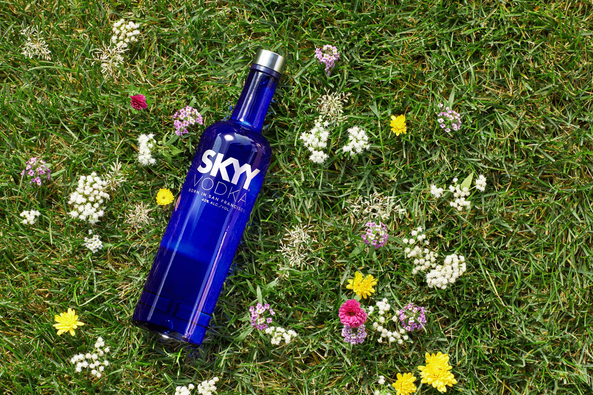 Drink 桌布 壁紙 — 一個充滿活力的藍色Skyy伏特加酒瓶放在綠草如茵、點綴著色彩繽紛野花的草地上。