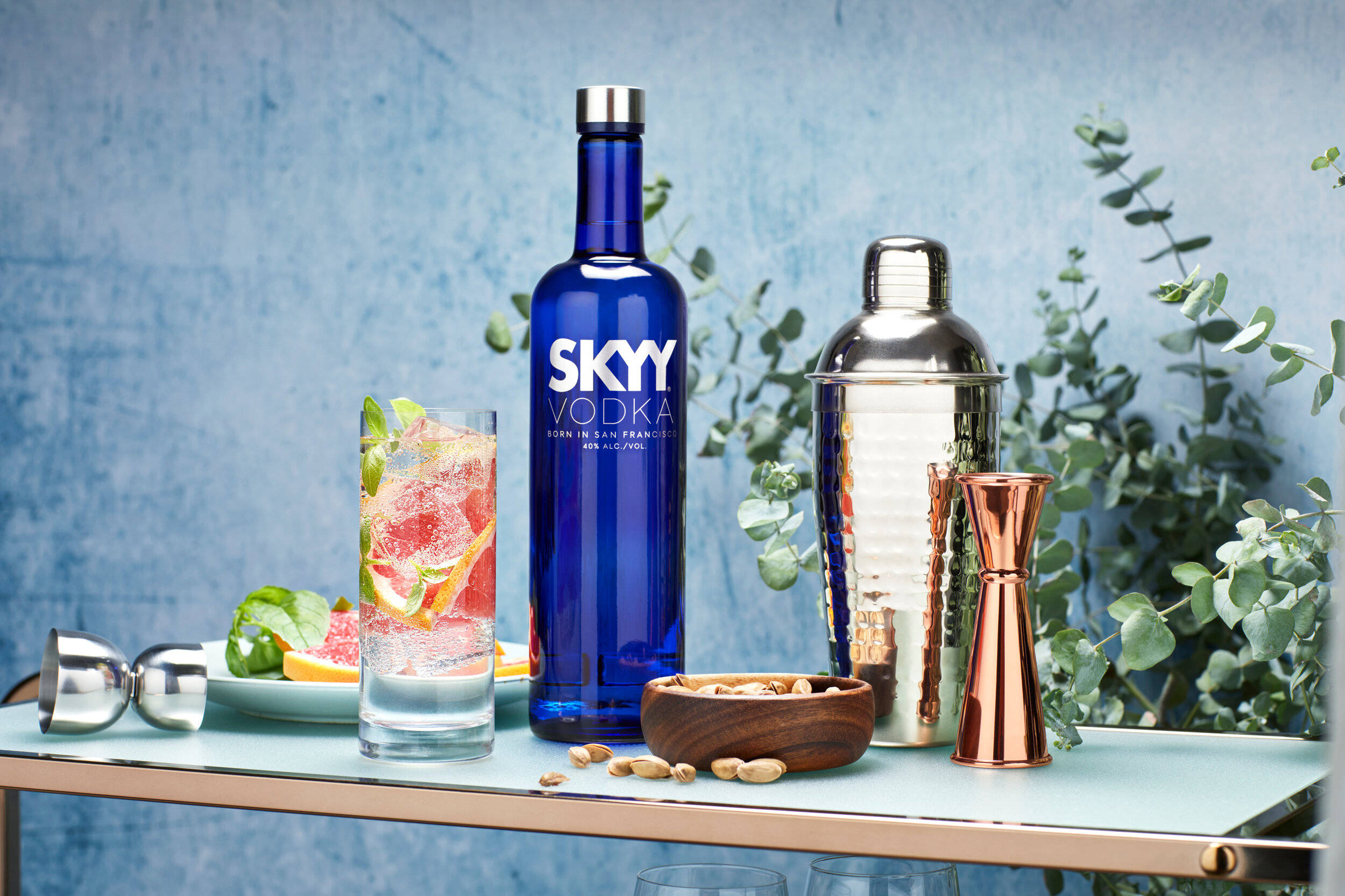 cocktail 桌布 壁紙 — 一個充滿活力的藍色Skyy伏特加酒瓶旁邊放著一個裝飾有柑橘和香草的雞尾酒杯，放在一個時尚的吧檯上，配有銅和銀的吧檯工具以及桉樹葉。