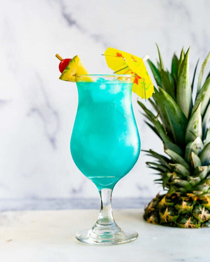 Blue Hawaii 桌布 壁紙 — 一個充滿活力的藍色熱帶雞尾酒，裝在颶風杯中，飾以鳳梨、櫻桃和傘，旁邊是大理石上的新鮮鳳梨。