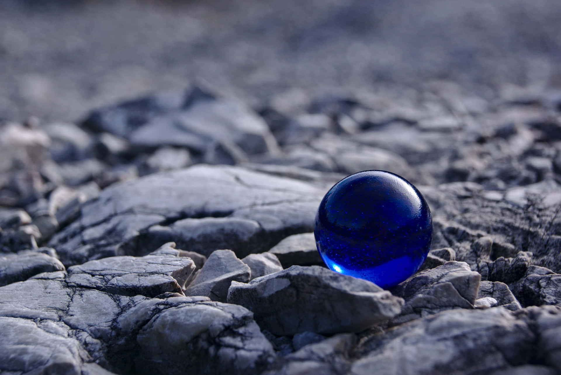 Blue Marble 桌布 壁紙 — 一個充滿活力的藍色玻璃球體擱在裂開的灰色岩石上，反射出光澤的光線。