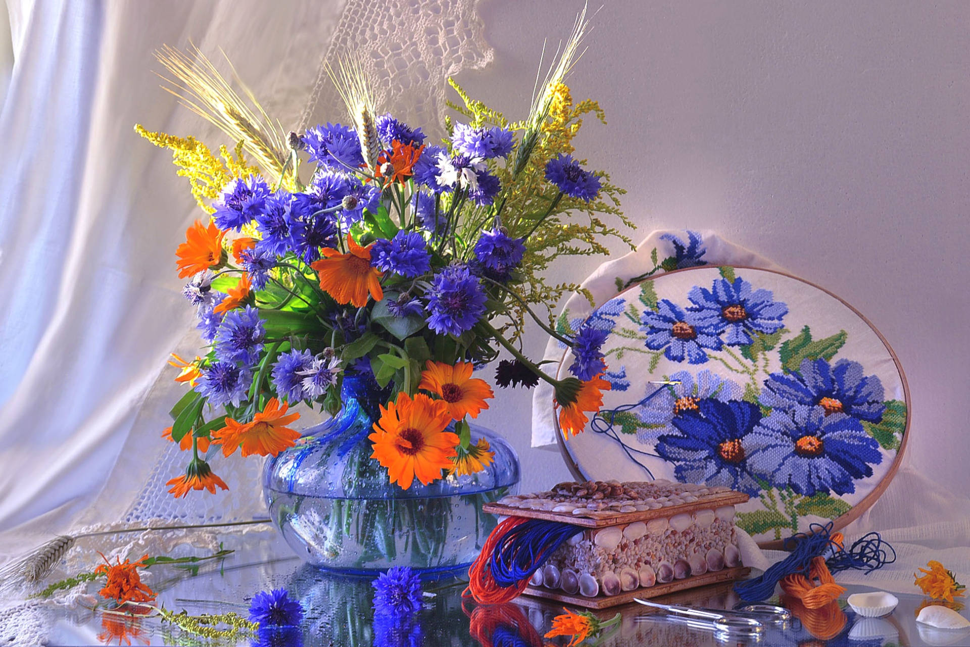 Flowers 桌布 壁紙 — 一個充滿活力的藍色玻璃花瓶盛載著紫色矢車菊和橙色萬壽菊，旁邊有一個帶有藍色花卉刺繡的繡花圈，以及散落在反光表面上的貝殼。