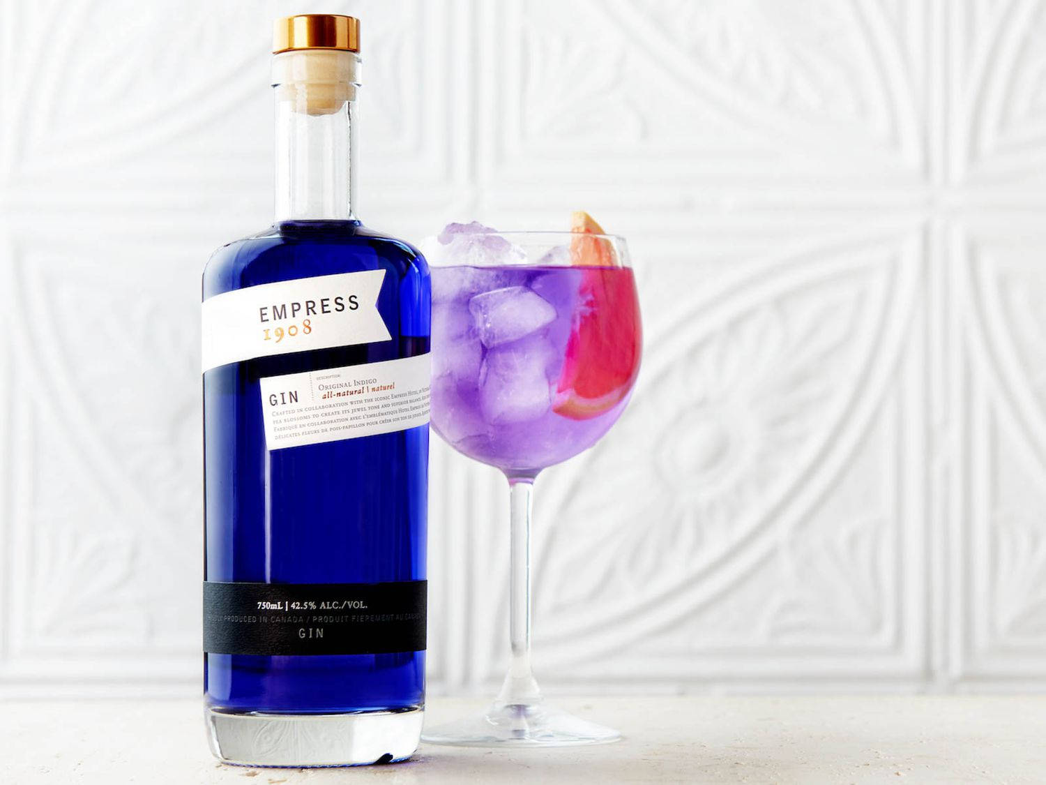 cocktail 桌布 壁紙 — 一個充滿活力的藍色琴酒瓶旁邊放著一杯以柑橘裝飾的紫色雞尾酒，背景是白色紋理牆。