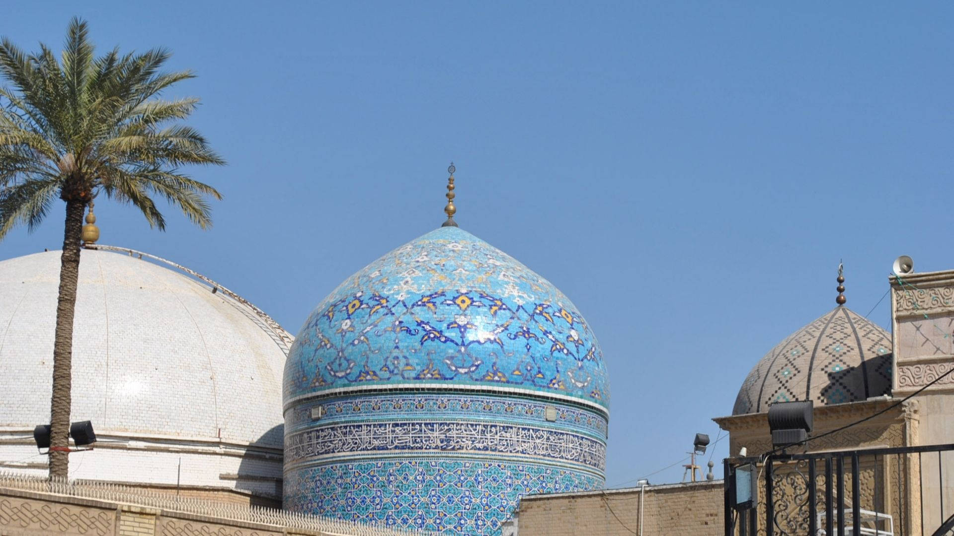 Baghdad 桌布 壁紙 — 一個充滿活力的藍色瓷磚圓頂，飾有精緻的花卉圖案，矗立在晴朗的天空下，旁邊有棕櫚樹和白色圓頂。