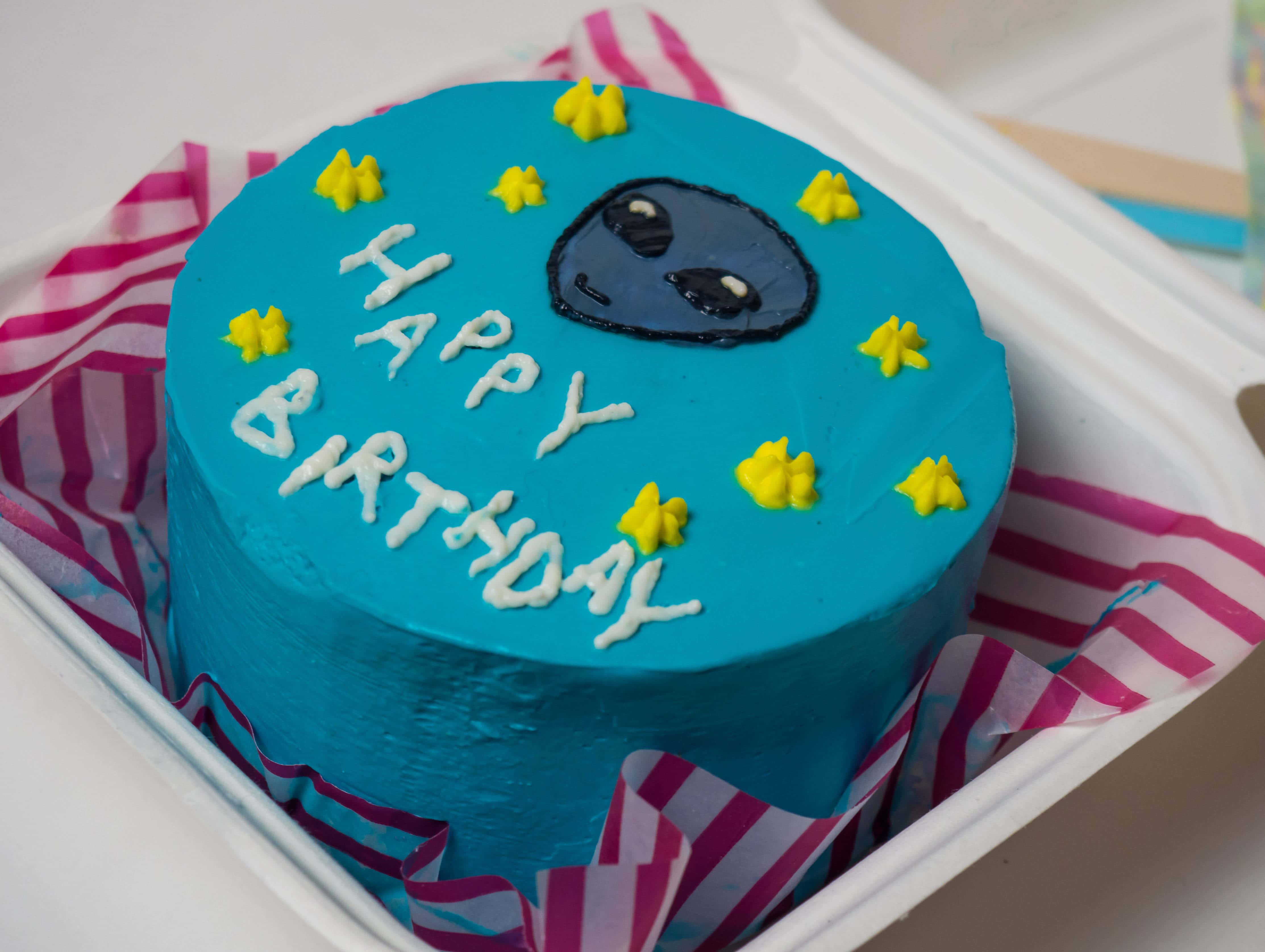 搞笑 桌布 壁紙 — 一個充滿活力的藍色生日蛋糕，上面有可愛的外星人臉和黃色星星，放在粉紅色條紋包裝紙中。
