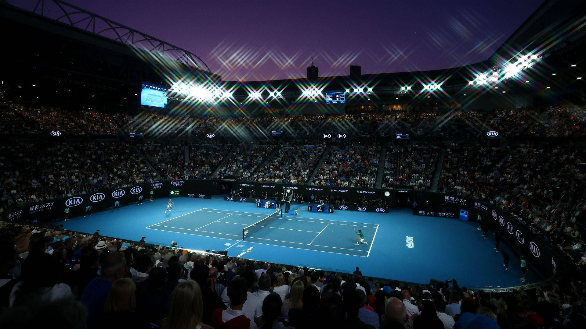 Australian Open 桌布 壁紙 — 一個充滿活力的藍色網球場在體育館燈光下閃耀，球員們在黃昏時分在座無虛席的觀眾面前比賽。