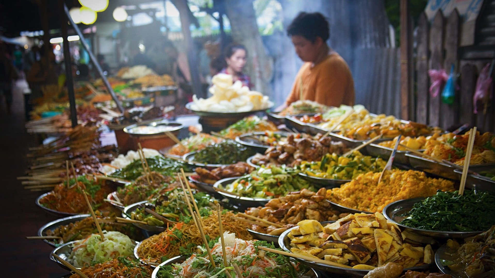 Indian 桌布 壁紙 — 一個充滿活力的街頭小吃攤，在溫暖的環境燈光下擺滿了色彩繽紛的菜餚和串燒。