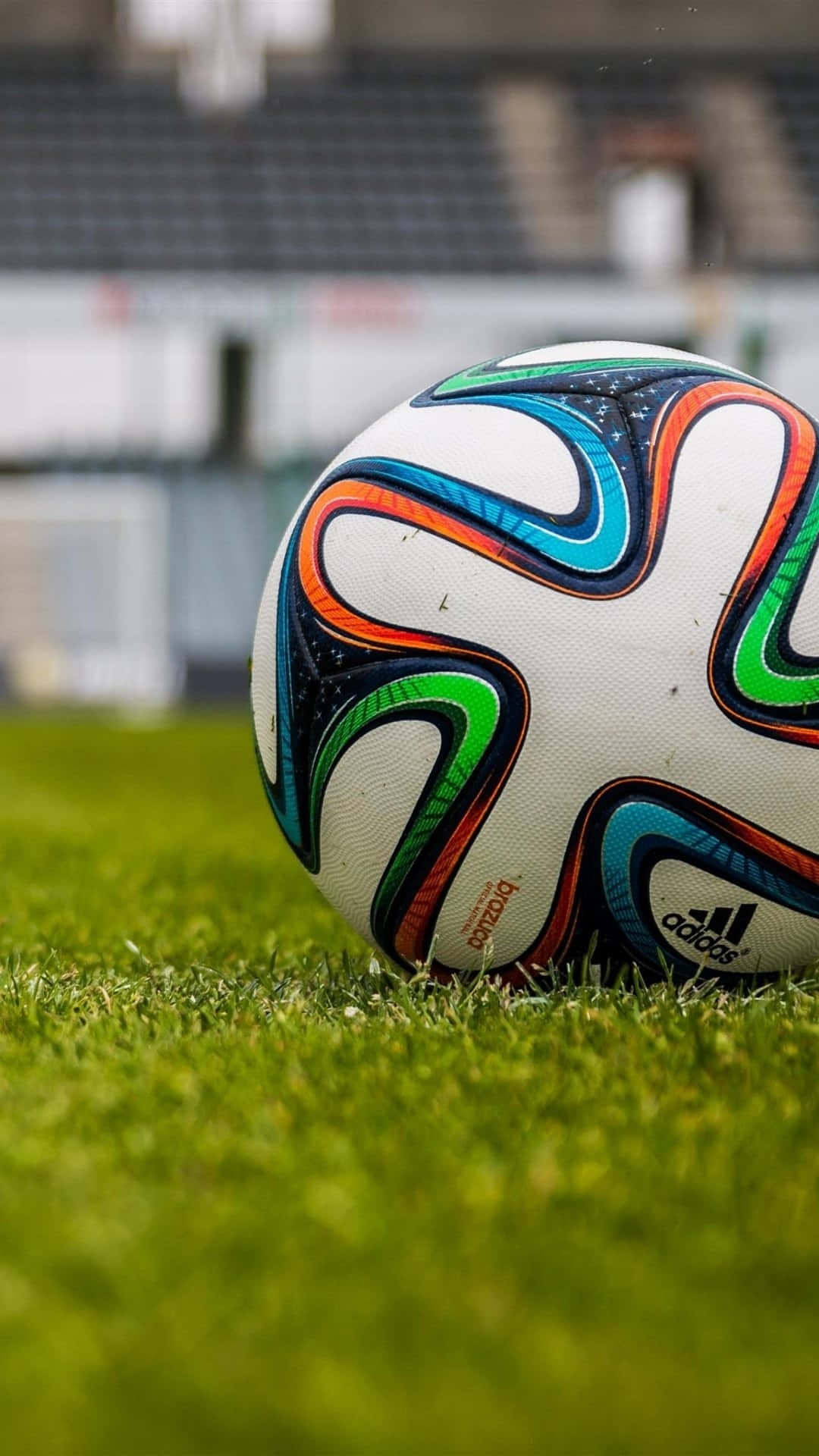 Soccer iPhone 桌布 壁紙 — 一個充滿活力的足球，帶有藍色、橙色和綠色的漩渦圖案，放置在體育館茂密的綠草地上。