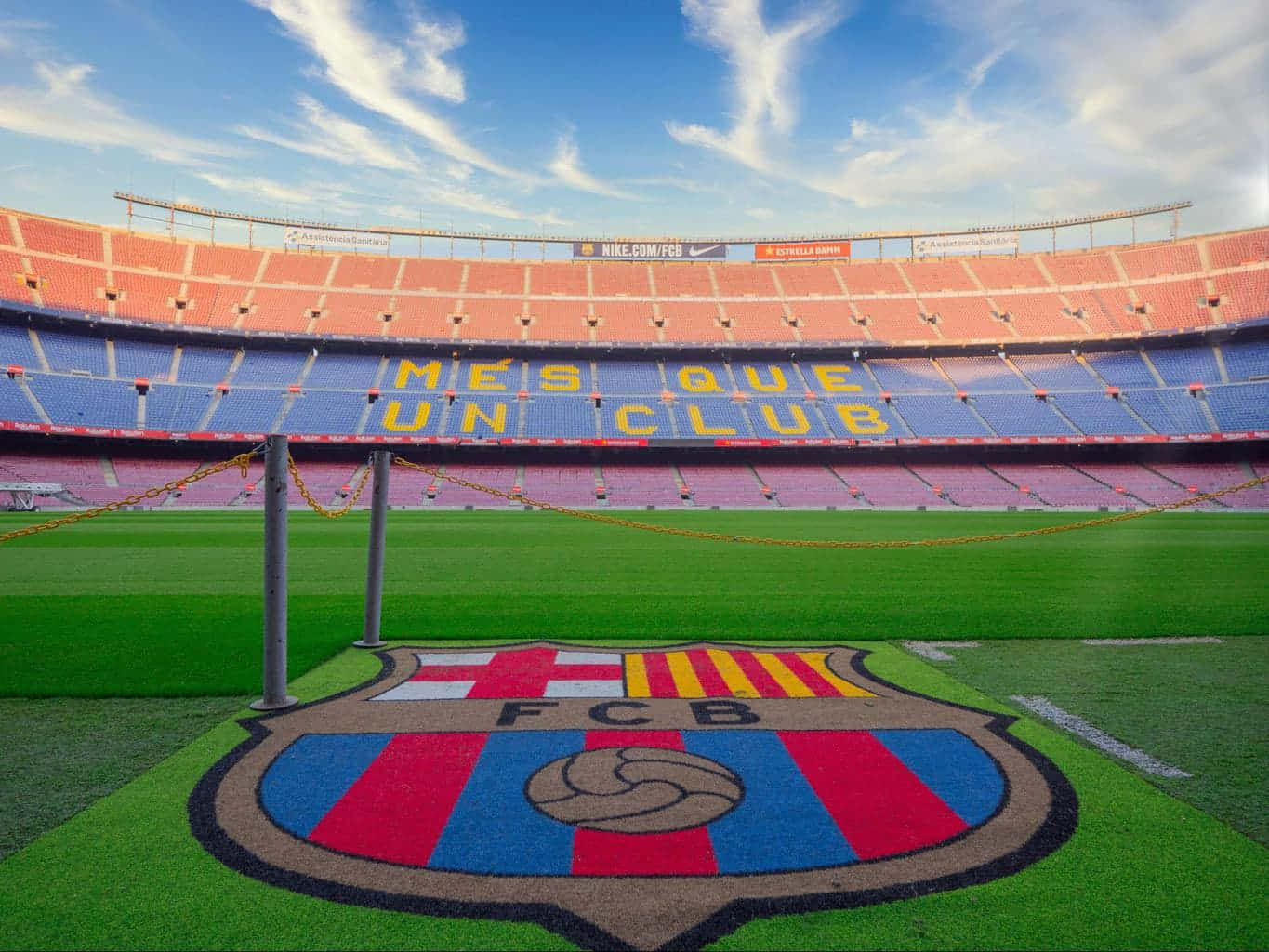 Barcelona 桌布 壁紙 — 一個充滿活力的足球場，球場上有大型FC巴塞隆納會徽，藍色座位上以黃色字母寫著「MES QUE UN CLUB」，上方是明亮的藍天。