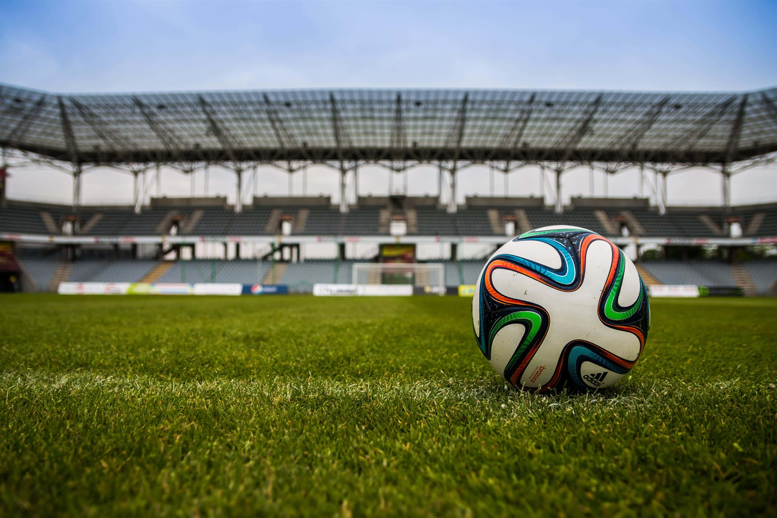 Soccer Ball 桌布 壁紙 — 一個充滿活力的足球放在空蕩蕩的體育館內茂密的綠草上，上方是清澈的藍天。
