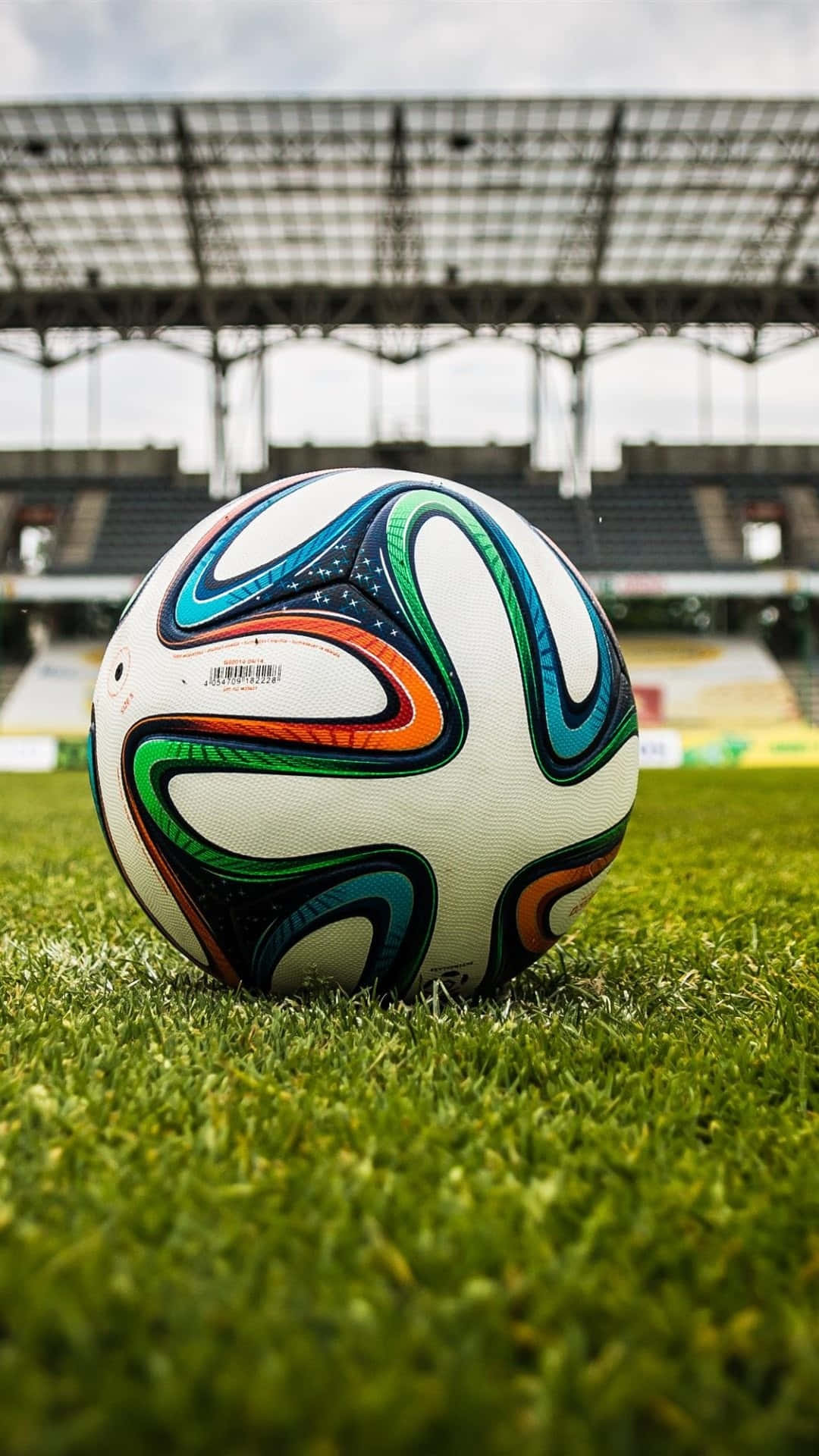 Soccer iPhone 桌布 壁紙 — 一個充滿活力的足球放在空蕩蕩的體育館內茂密的綠草上，天空多雲。