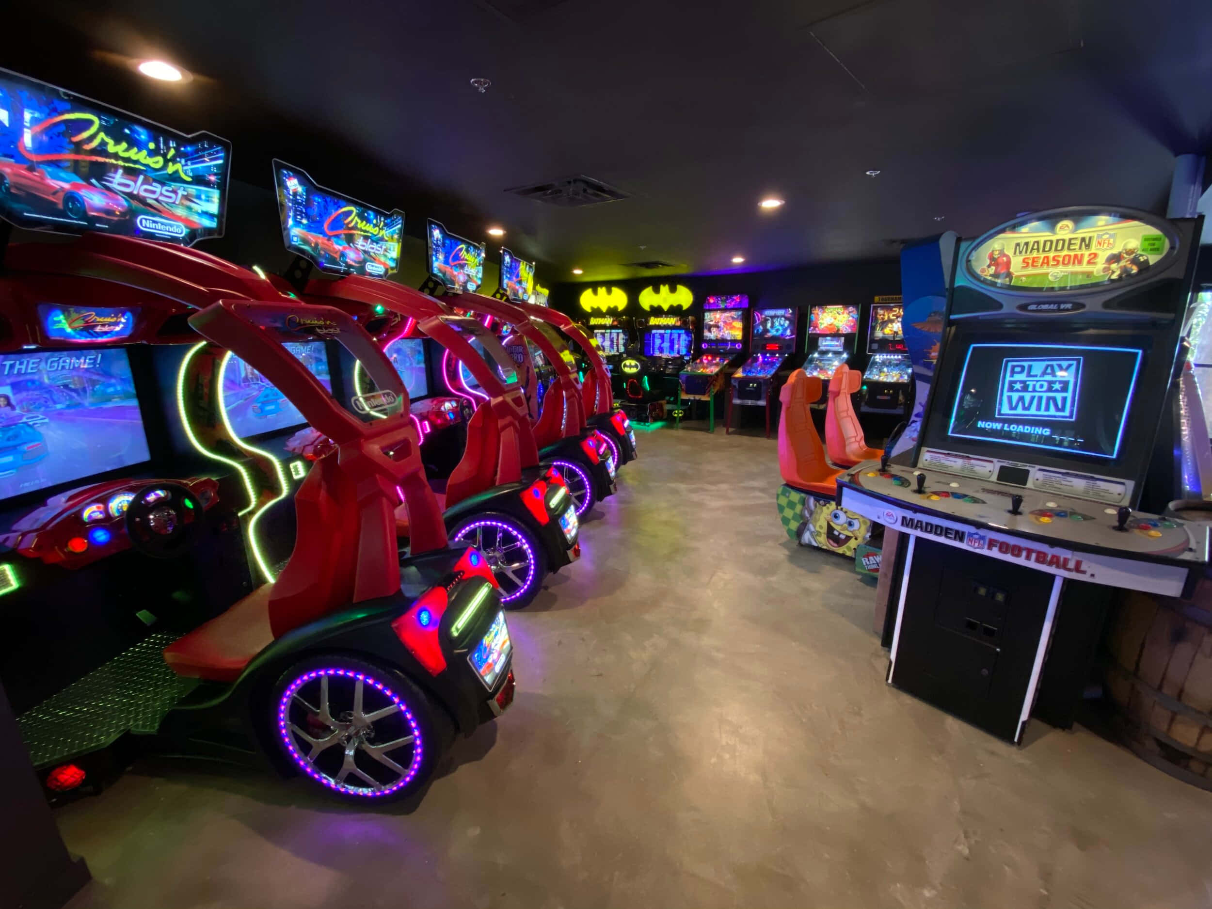 Arcade Games 桌布 壁紙 — 一個充滿活力的遊戲機中心，在色彩繽紛的霓虹燈下，擺滿了發光的賽車模擬器和Madden Football遊戲機。