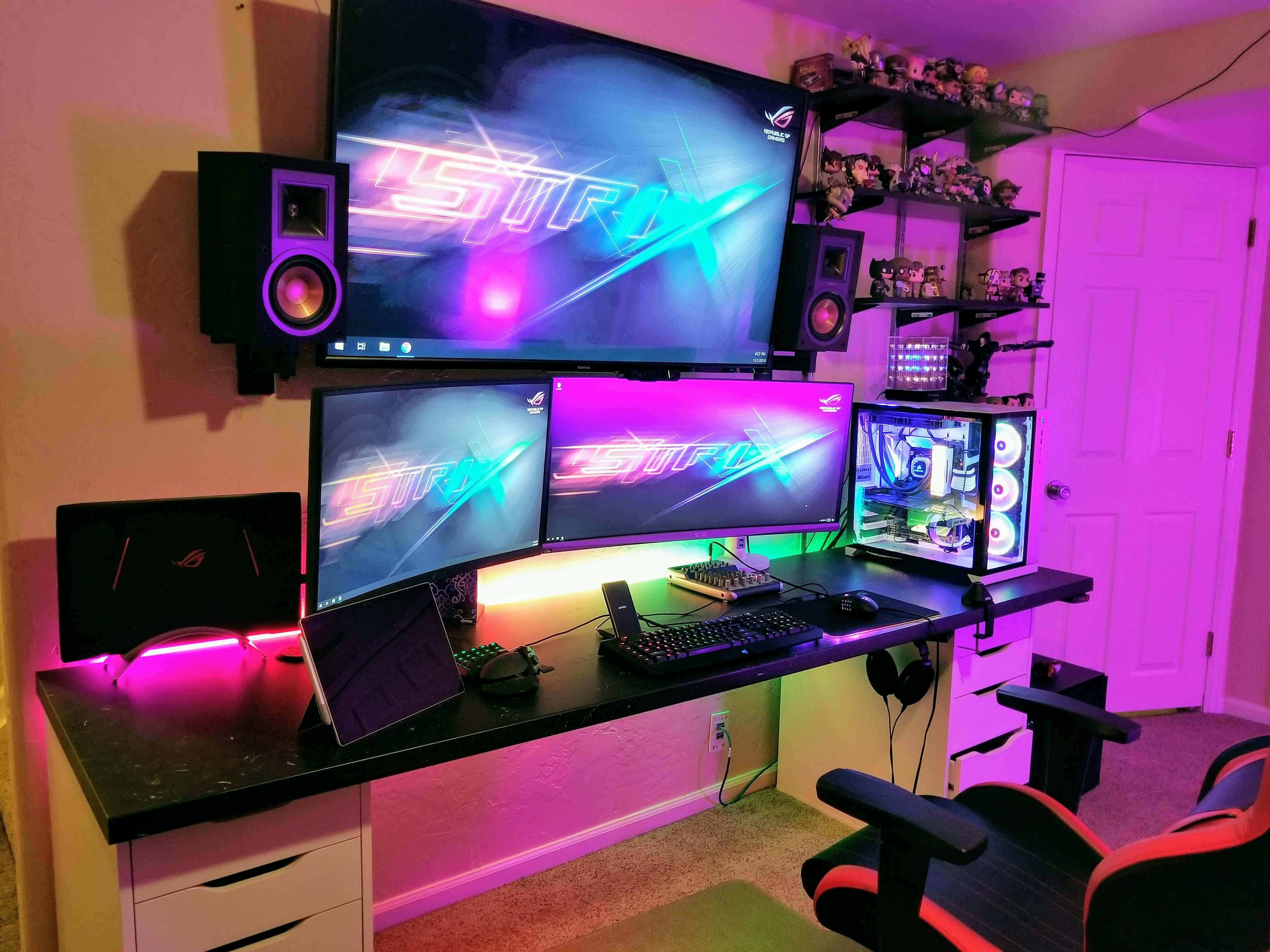 Gaming Pc Setup 桌布 壁紙 — 一個充滿活力的遊戲設置，多個顯示器發出紫色和藍色光芒，周圍環繞著RGB燈光和架子上的收藏人偶。