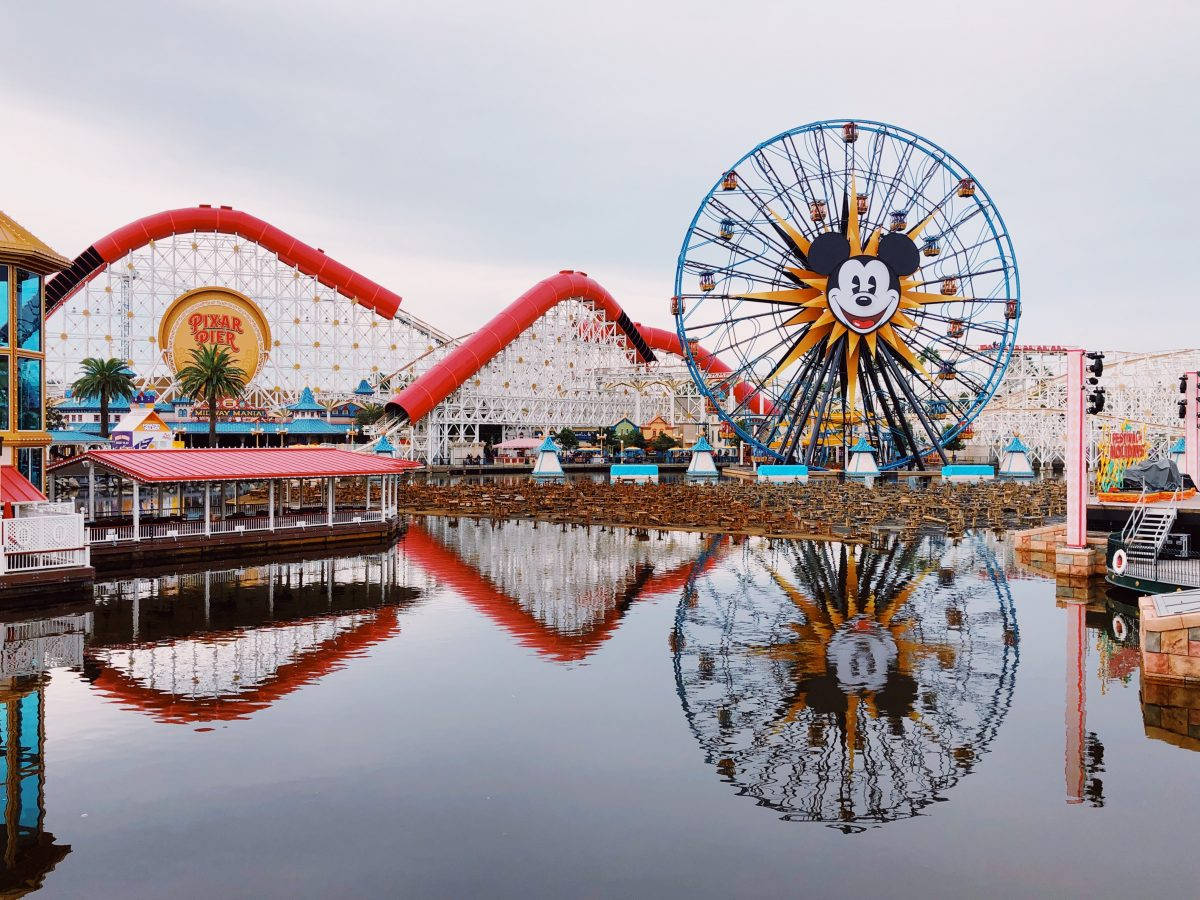 Disneyland 桌布 壁紙 — 一個充滿活力的遊樂園，有紅色雲霄飛車和米奇老鼠摩天輪，在陰天下的平靜水面上倒映。
