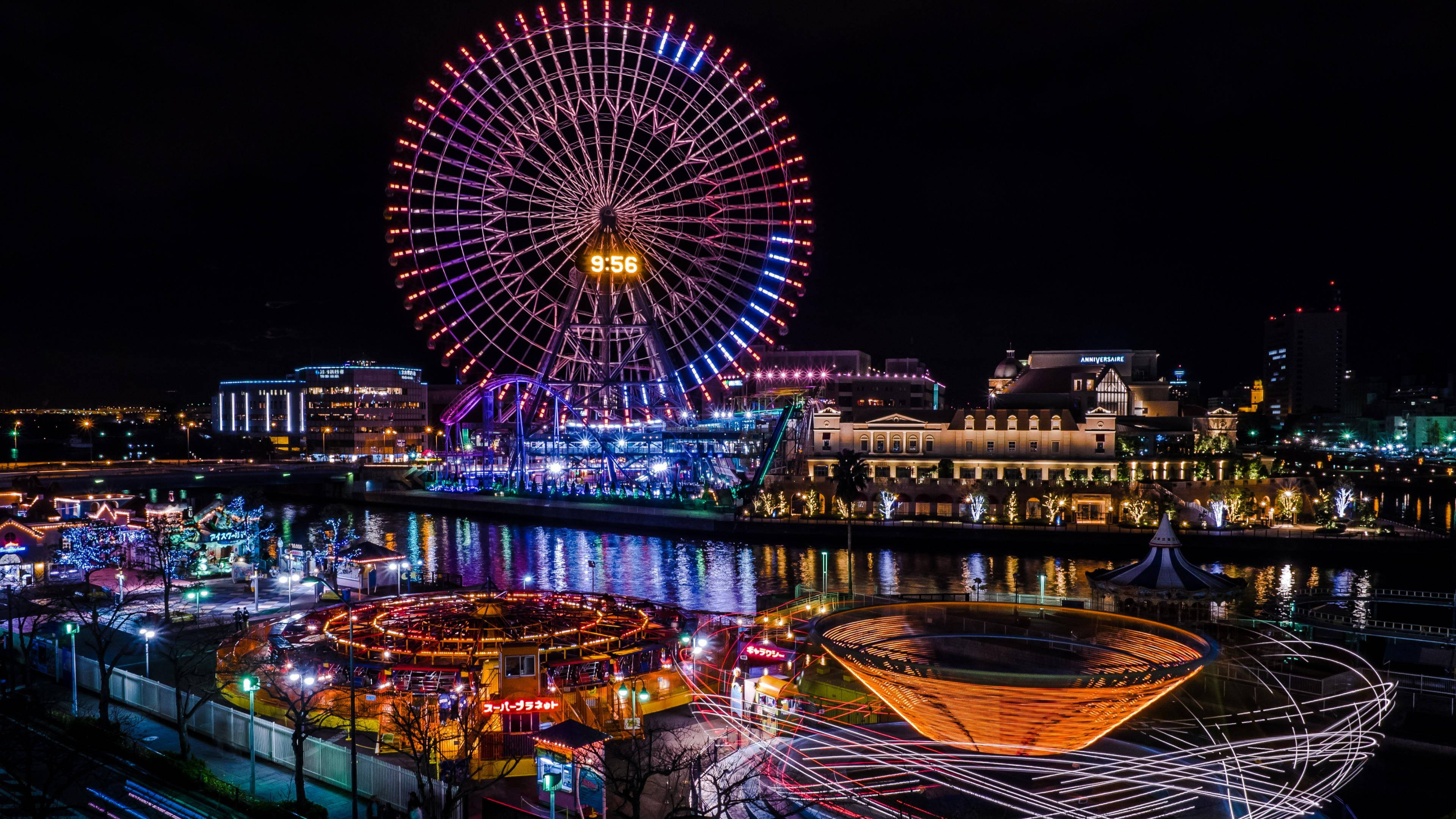 Theme Park 桌布 壁紙 — 一個充滿活力的遊樂園的耀眼夜景，巨大的發光摩天輪倒映在平靜的河面上。