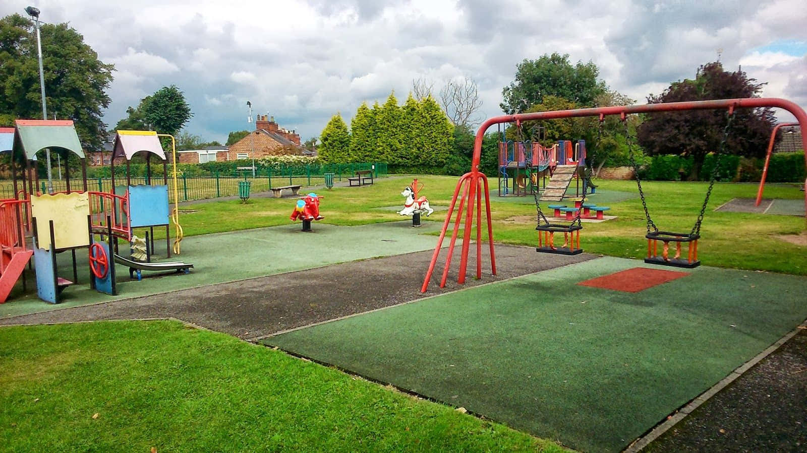 Playground 桌布 壁紙 — 一個充滿活力的遊樂場，設有紅色鞦韆和色彩繽紛的遊樂設施，背景是陰天和綠草。