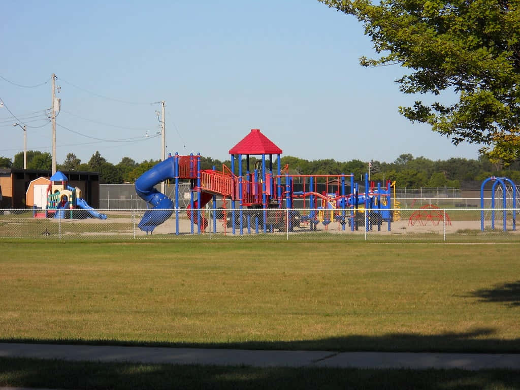 Playground 桌布 壁紙 — 一個充滿活力的遊樂場，設有藍色溜滑梯和紅色屋頂，矗立在晴朗藍天下的草地上。