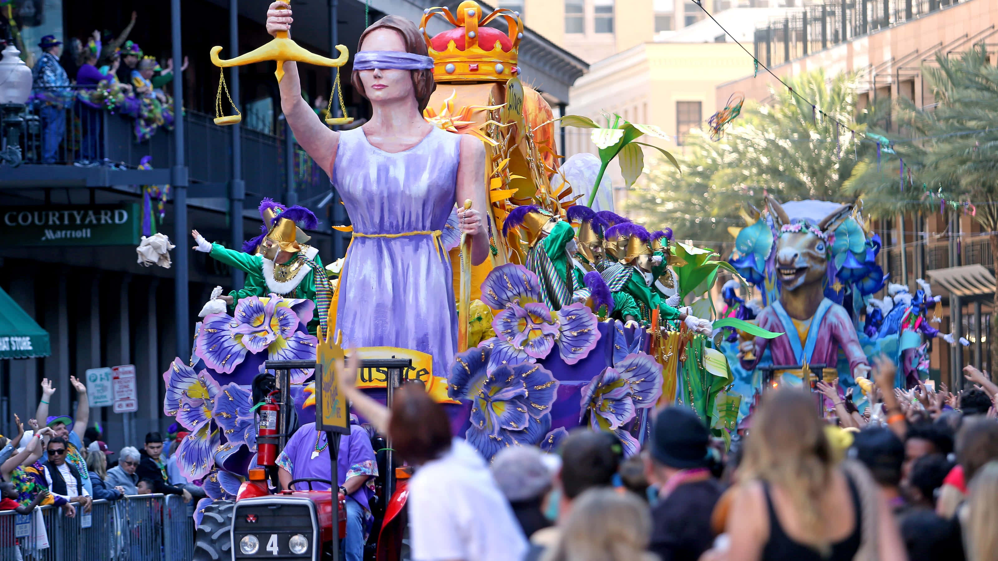 Mardi Gras 桌布 壁紙 — 一個充滿活力的遊行花車，展示了一位蒙眼的正義女神，手持天平，周圍是色彩繽紛的花卉裝飾和歡呼的人群。