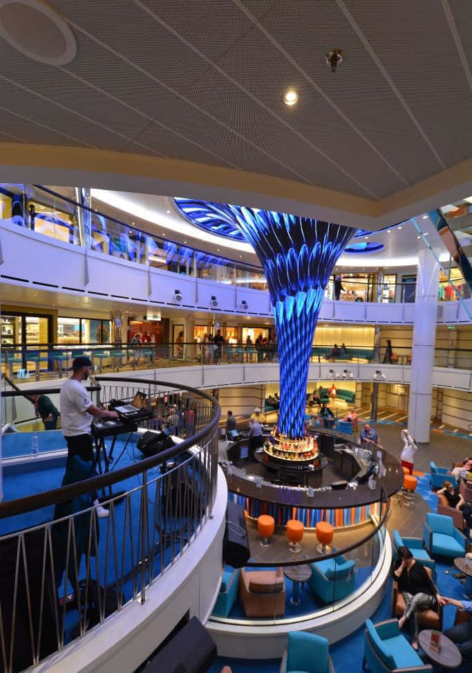 Carnival Vista 桌布 壁紙 — 一個充滿活力的郵輪中庭，設有發光的藍色中央雕塑，人們在陽台和座位區放鬆。