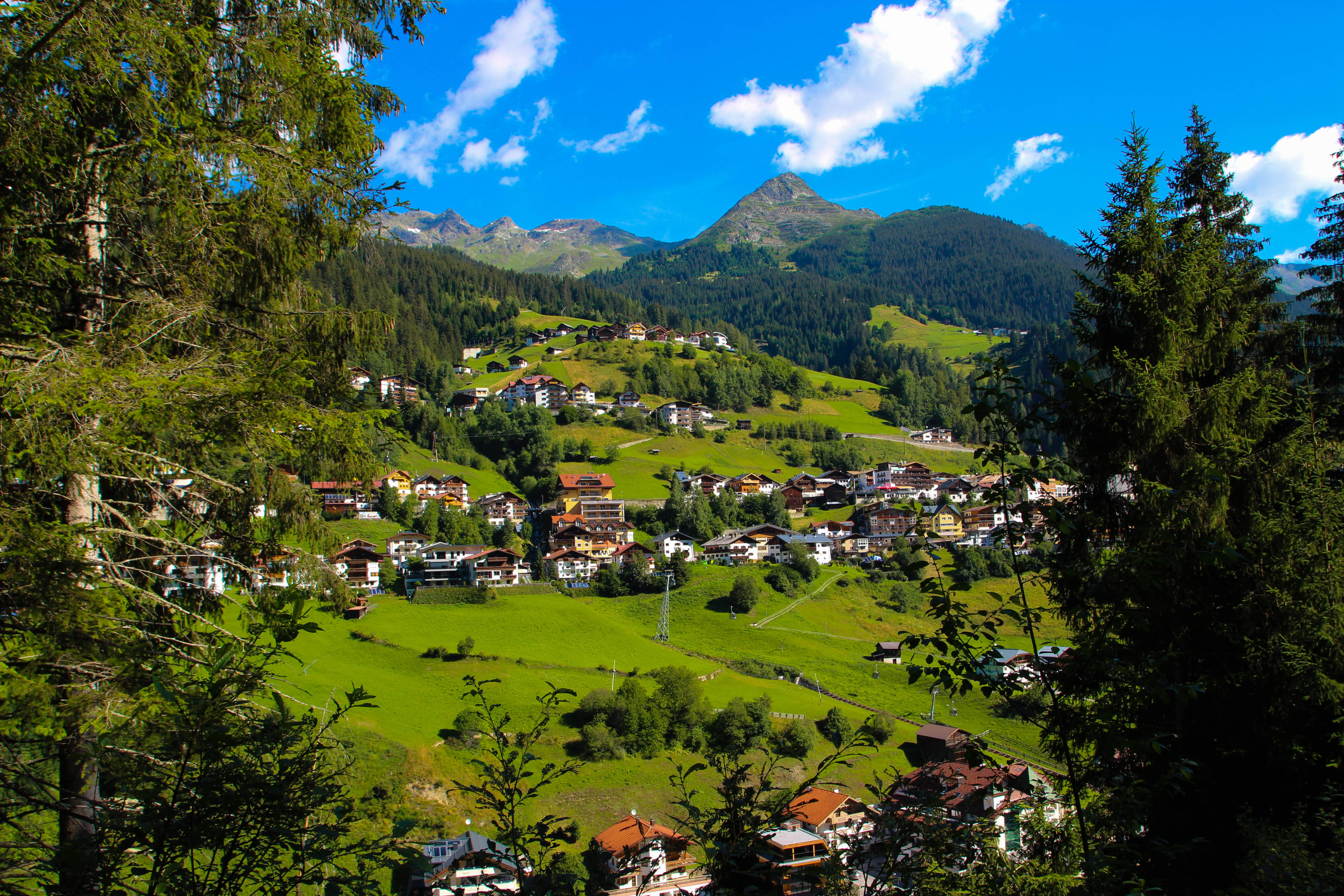 Austria 桌布 壁紙 — 一個充滿活力的阿爾卑斯山村莊坐落在鬱鬱蔥蔥的綠色山谷中，周圍環繞著高聳的松樹和白雪覆蓋的山脈，在明亮的藍天下。